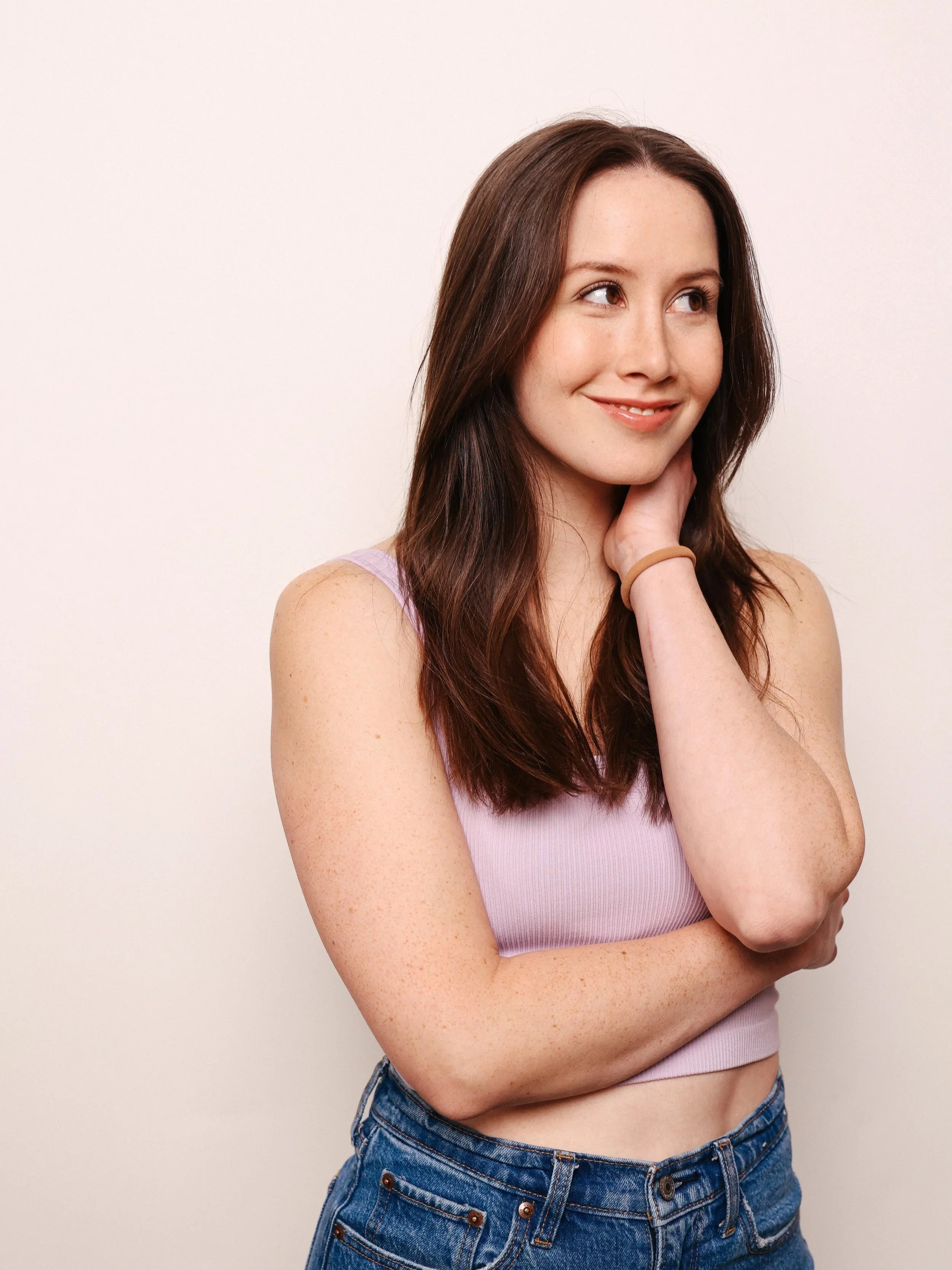 A young woman with long brown hair smiling, wearing a lavender crop top and blue jeans, standing against a plain light-colored background.