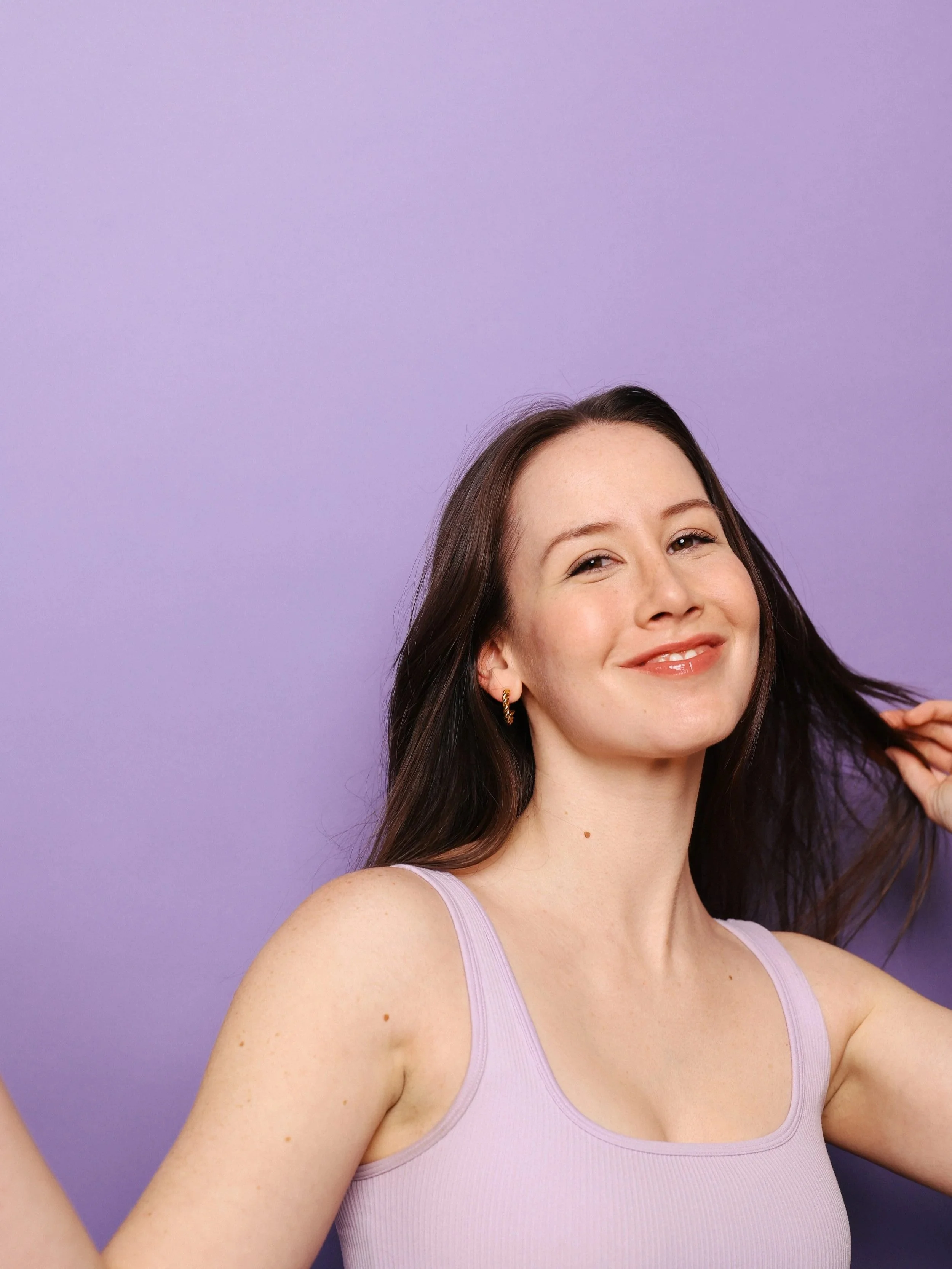 A young woman with long brown hair, smiling with a slight tilt of her head, wearing a lavender tank top, standing against a purple background.