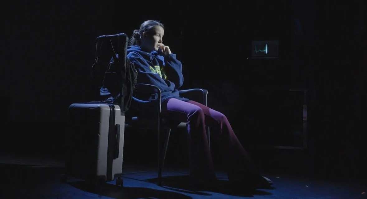 A young woman sitting alone in a dark room, wearing a hoodie, with luggage beside her, appearing to be waiting or pondering during a medical examination or emergency situation.