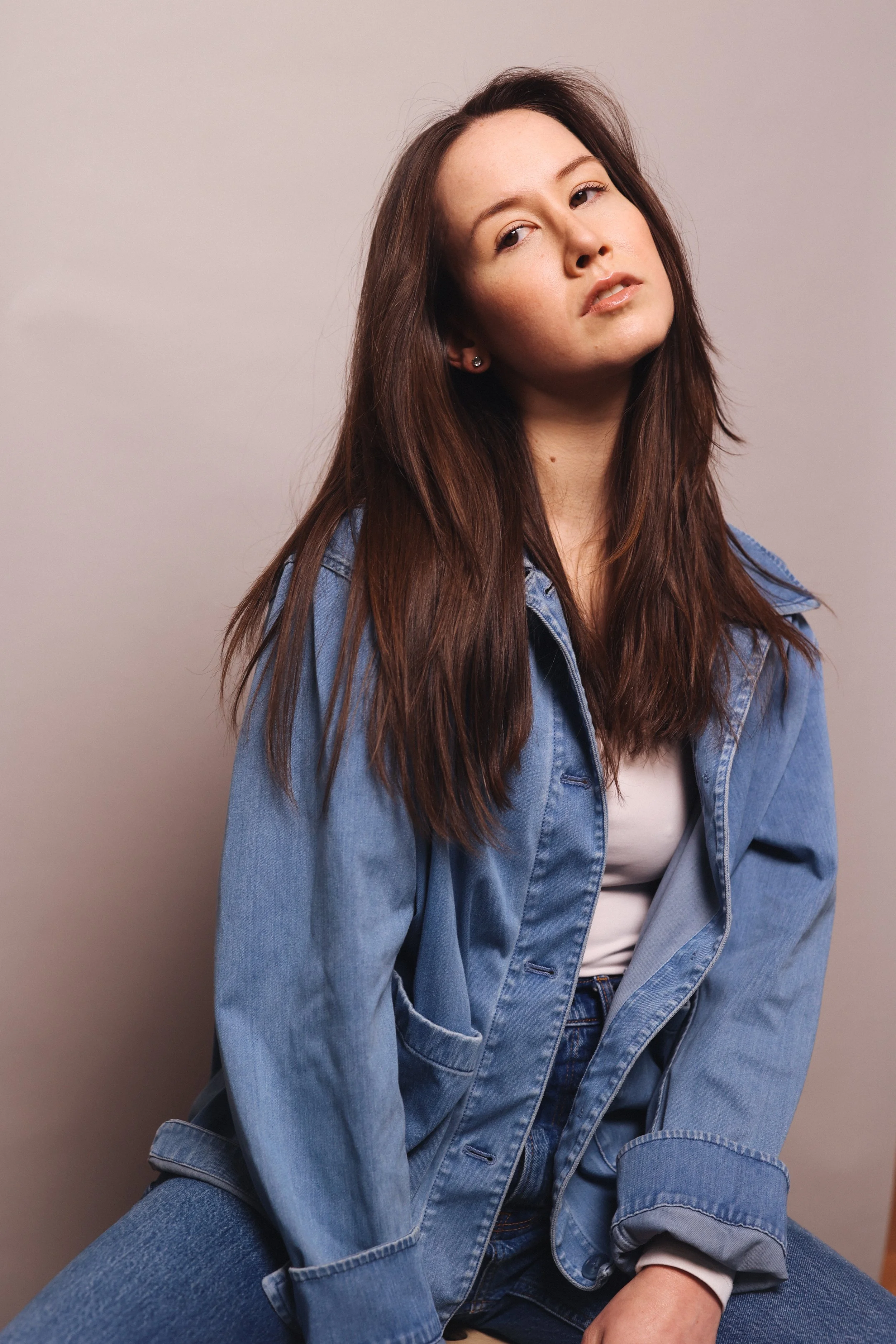 A young woman with long brown hair wearing a denim jacket and jeans, seated against a plain wall, looking slightly to the side.