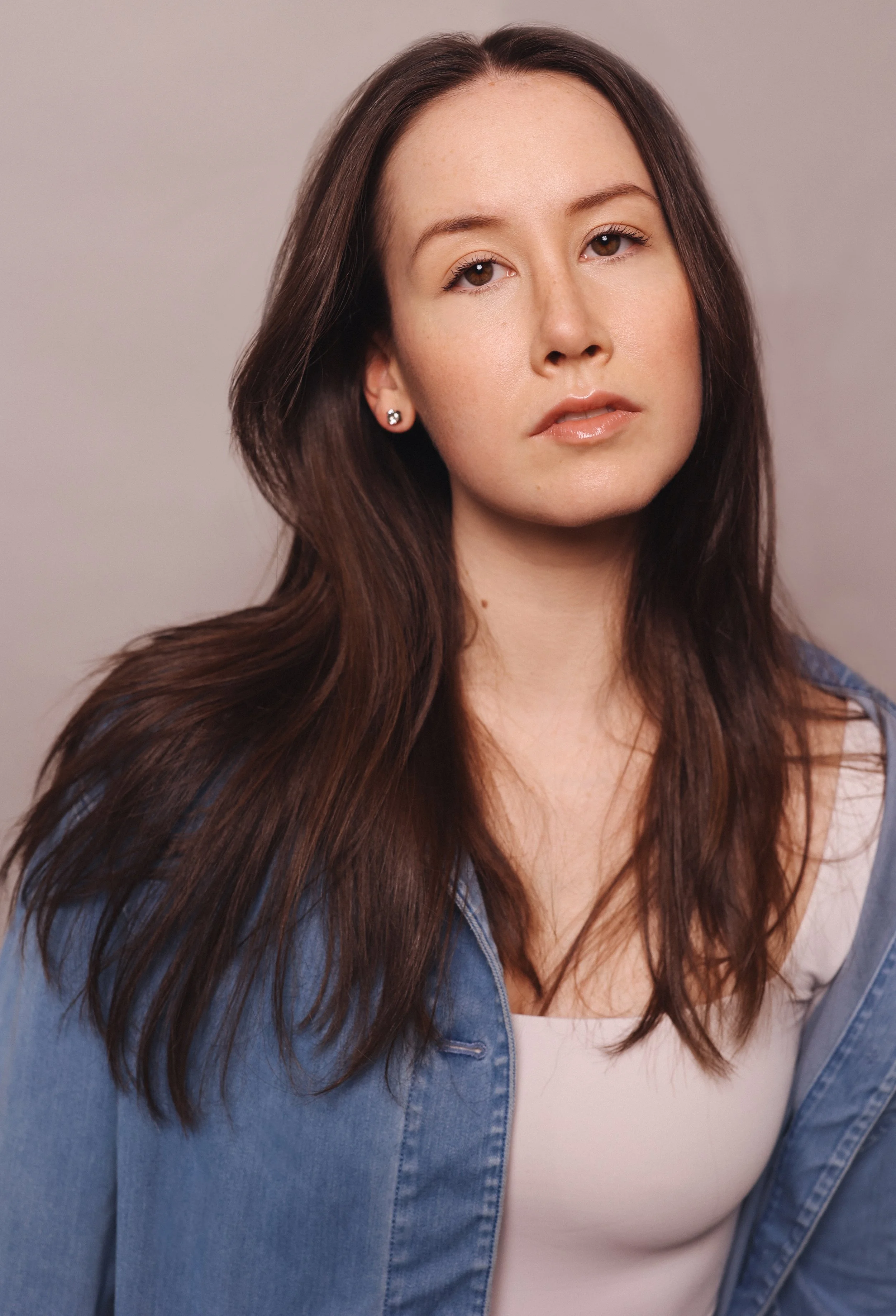 Portrait of a young woman with long brown hair, wearing a denim jacket and a white top, looking directly at the camera with a neutral expression.