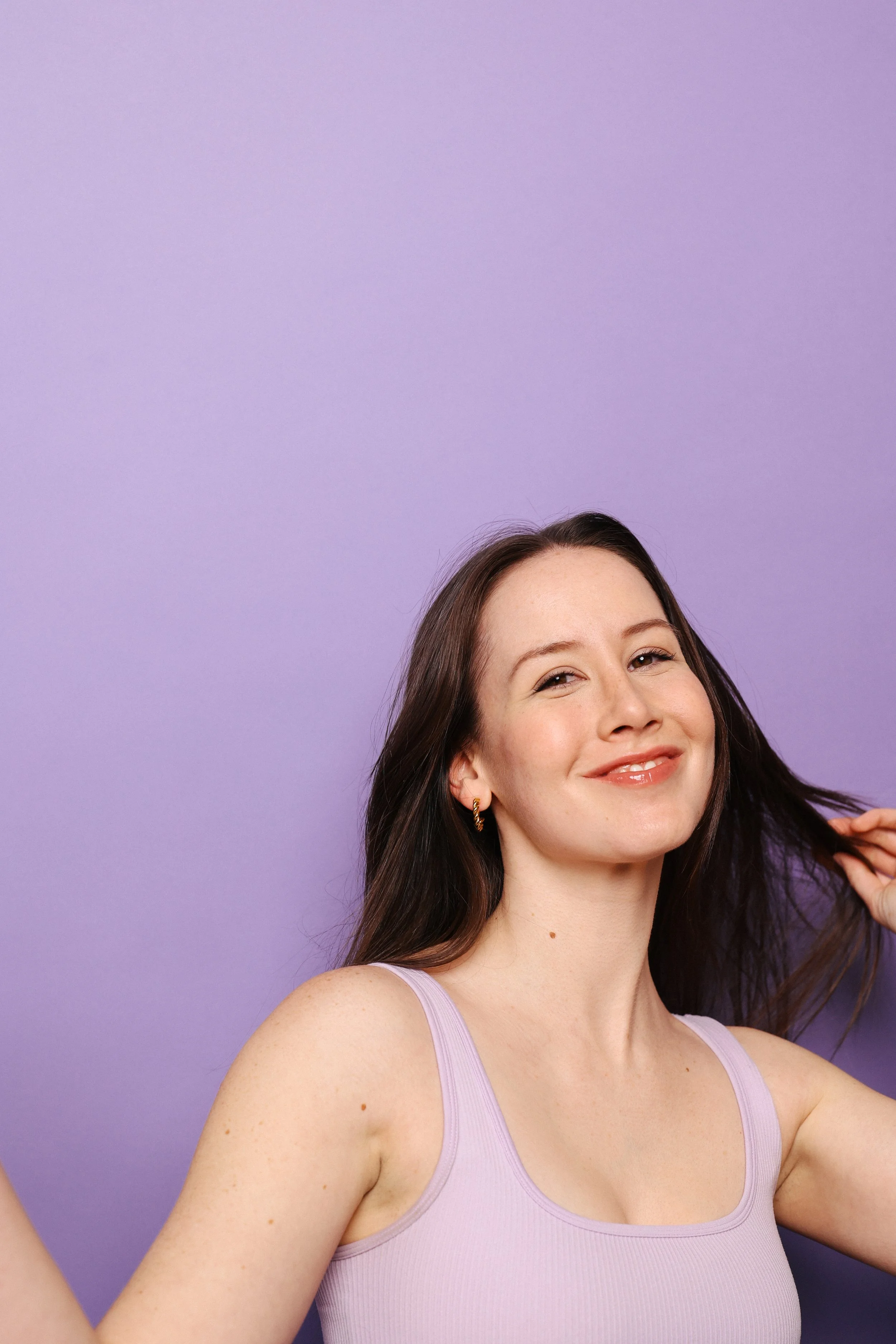 A young woman with long brown hair, smiling with a slight tilt of her head, wearing a lavender tank top, standing against a purple background.