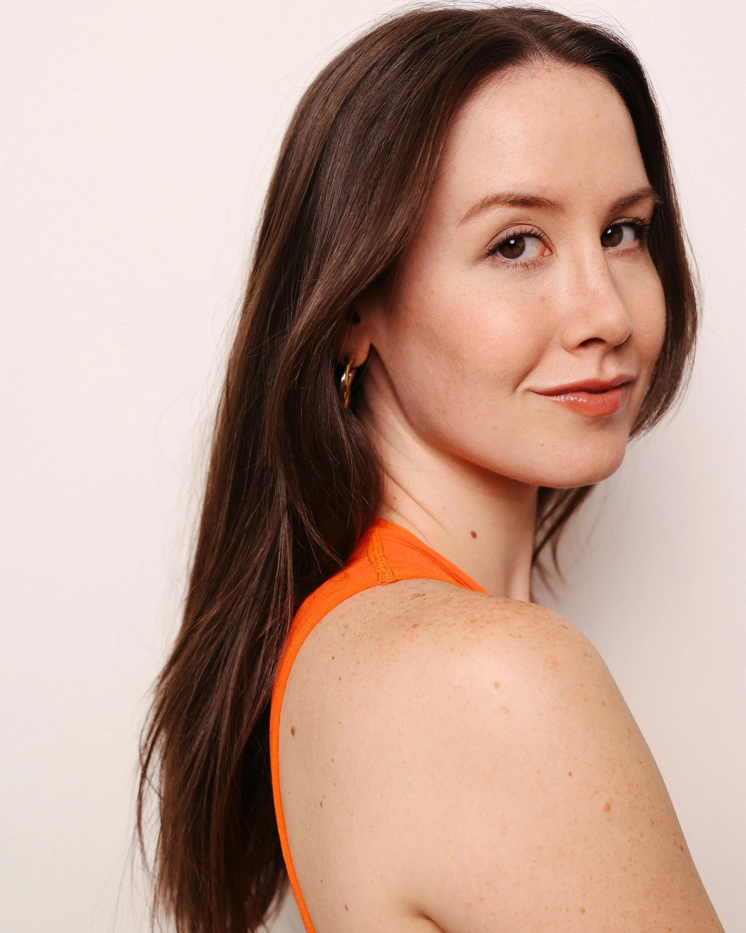A woman with long brown hair and light skin, wearing an orange sleeveless top and gold hoop earrings, smiling softly against a plain white background.