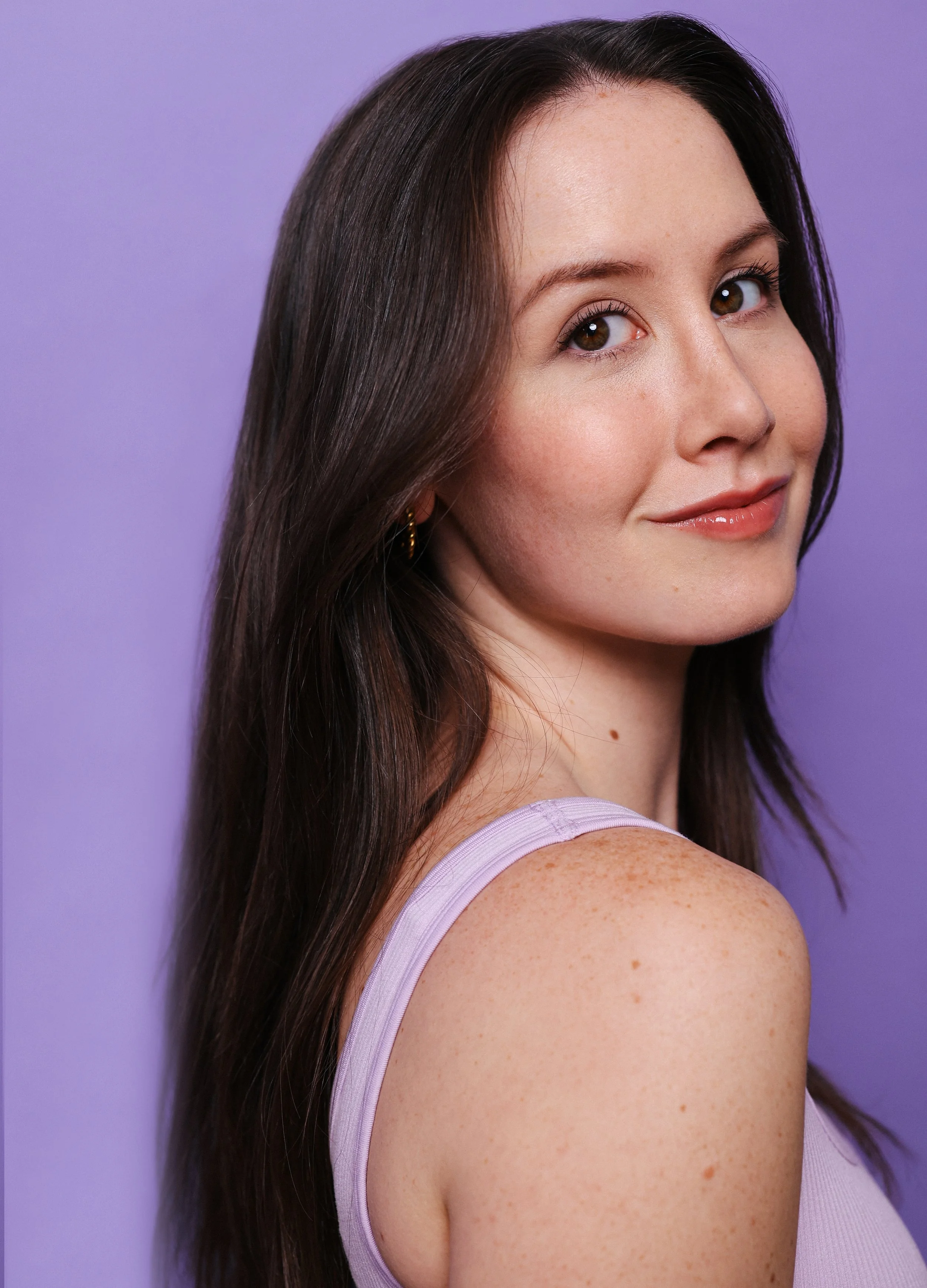 Portrait of a young woman with long brown hair, smiling slightly, wearing a lavender sleeveless top, against a purple background.