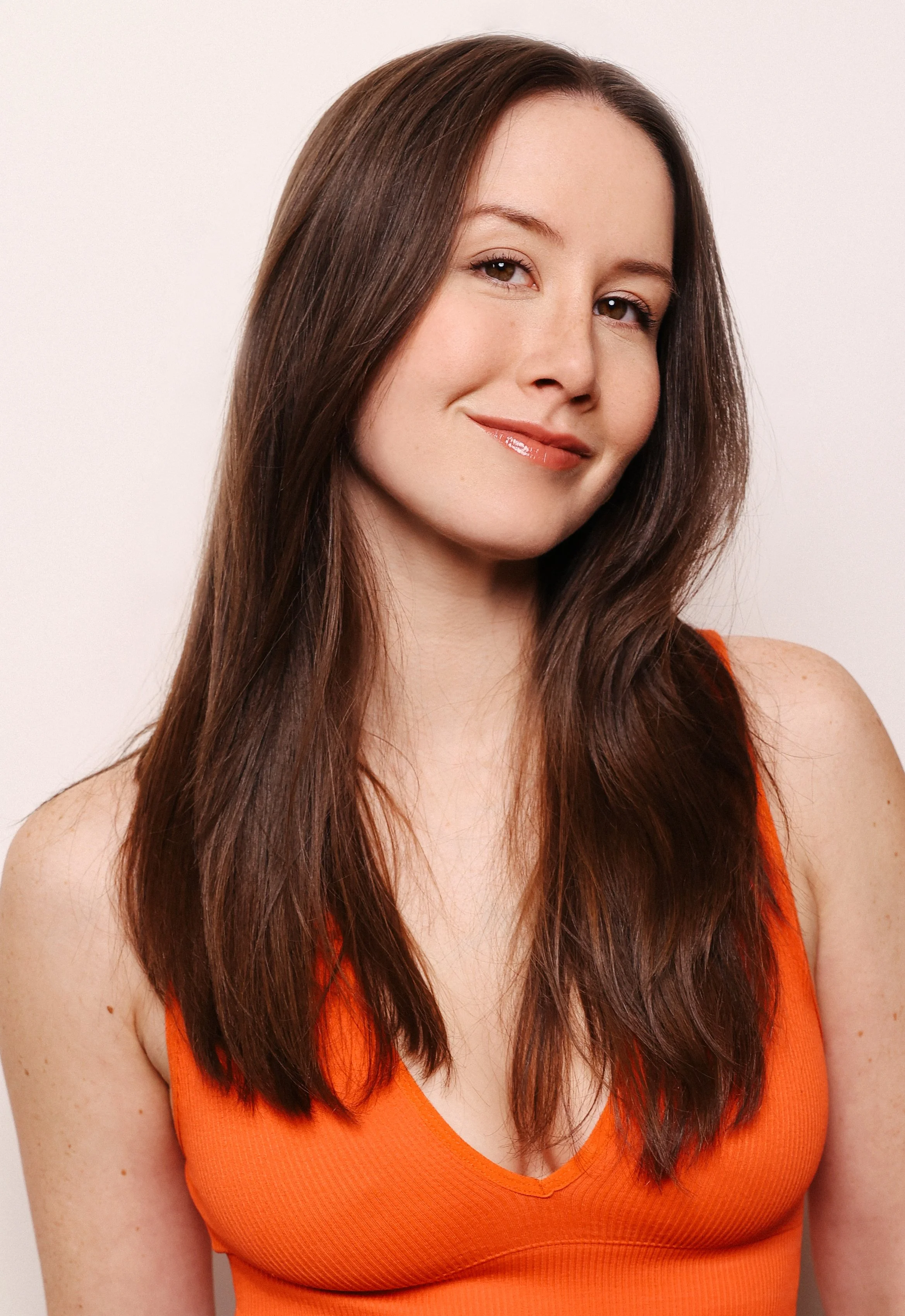 A woman with long brown hair, light skin, wearing an orange sleeveless top, smiling slightly against a plain white background.