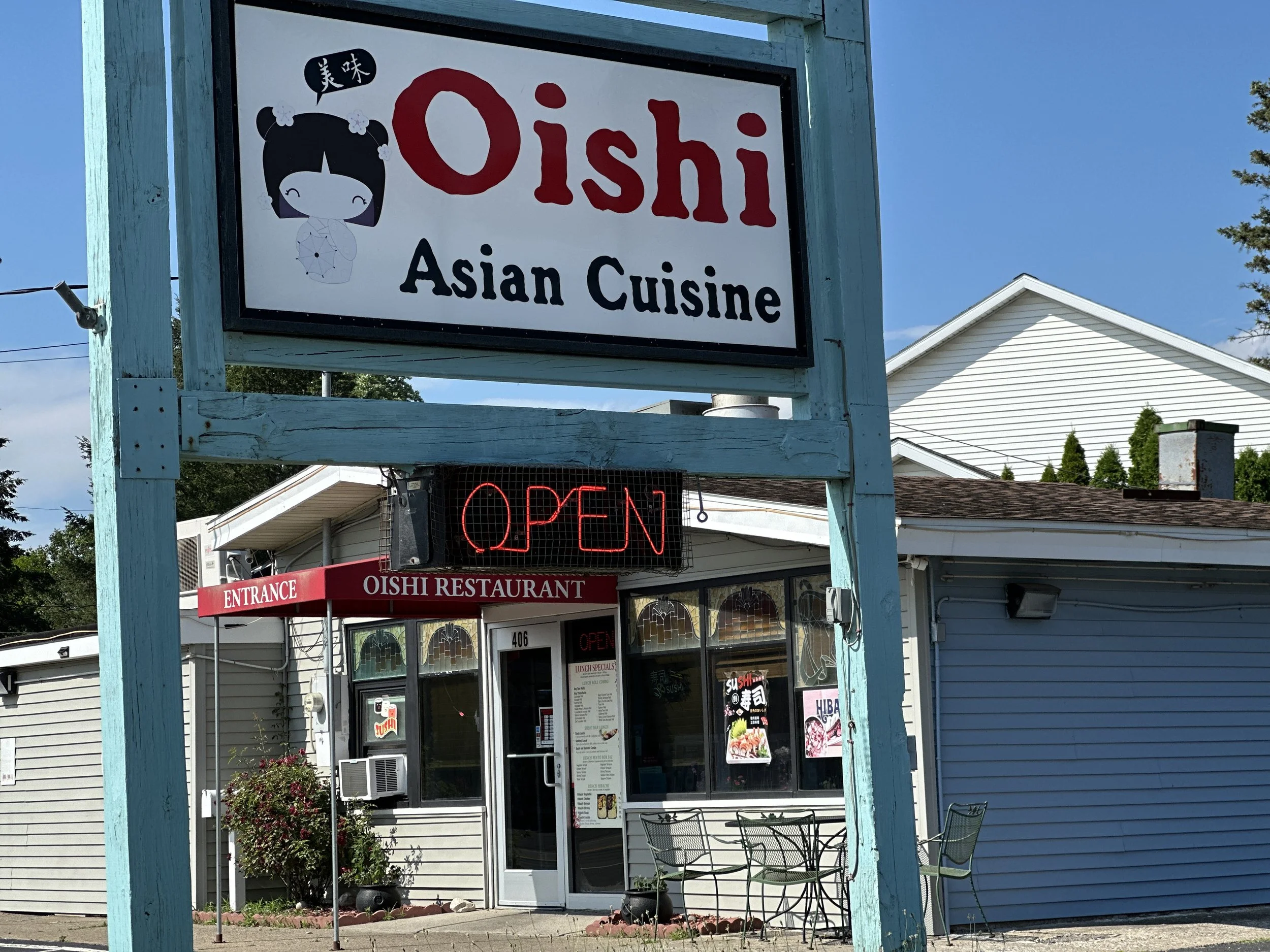 Sign for Oishi Asian Cuisine restaurant with an 'Open' sign in neon, storefront with tables and chairs outside, and a white building in the background.