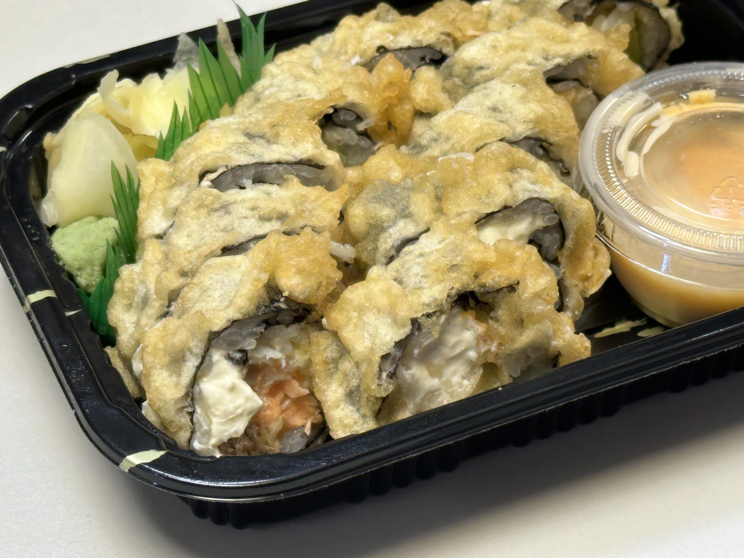 A black tray of tempura sushi rolls topped with crispy tempura batter, served with wasabi, pickled ginger, and a container of dipping sauce.