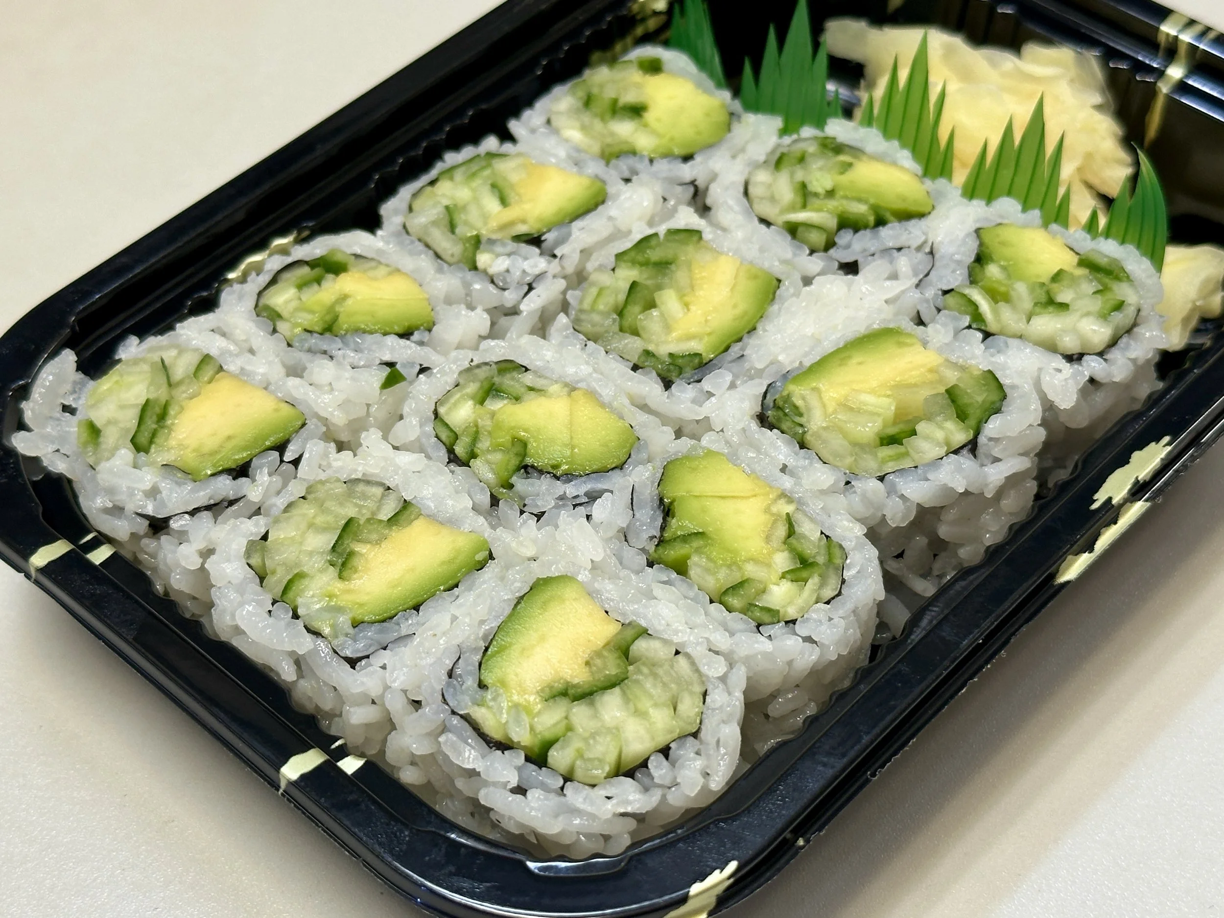 A black plastic takeout container filled with sushi rolls topped with sliced cucumber. There are pieces of tempura and green plastic divider inside.