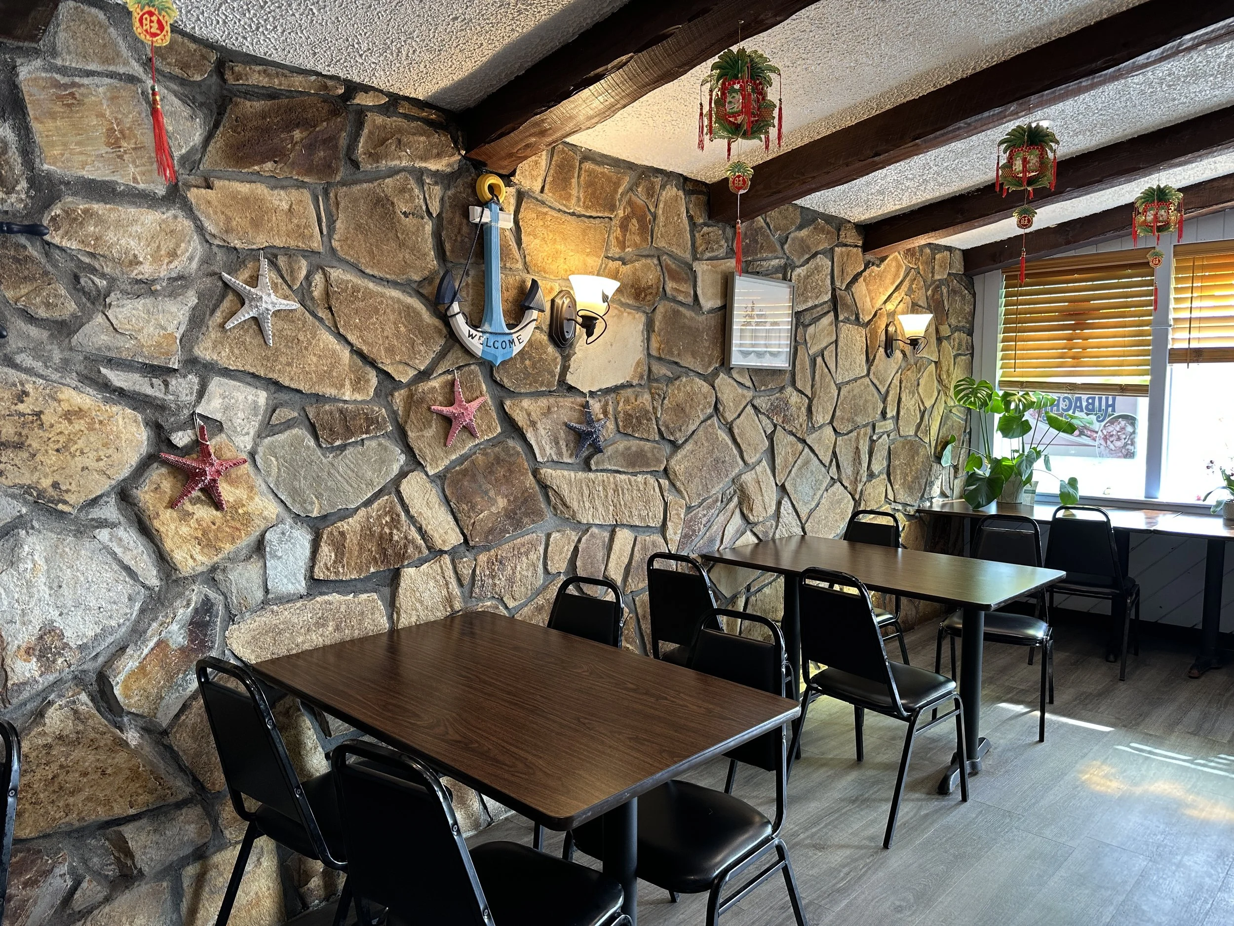 Interior of a restaurant or cafe with a stone wall decorated with starfish and nautical items, hanging plants, and wooden beams on the ceiling. There are rectangular tables with black chairs, a window with blinds, and potted plants on the windowsill.