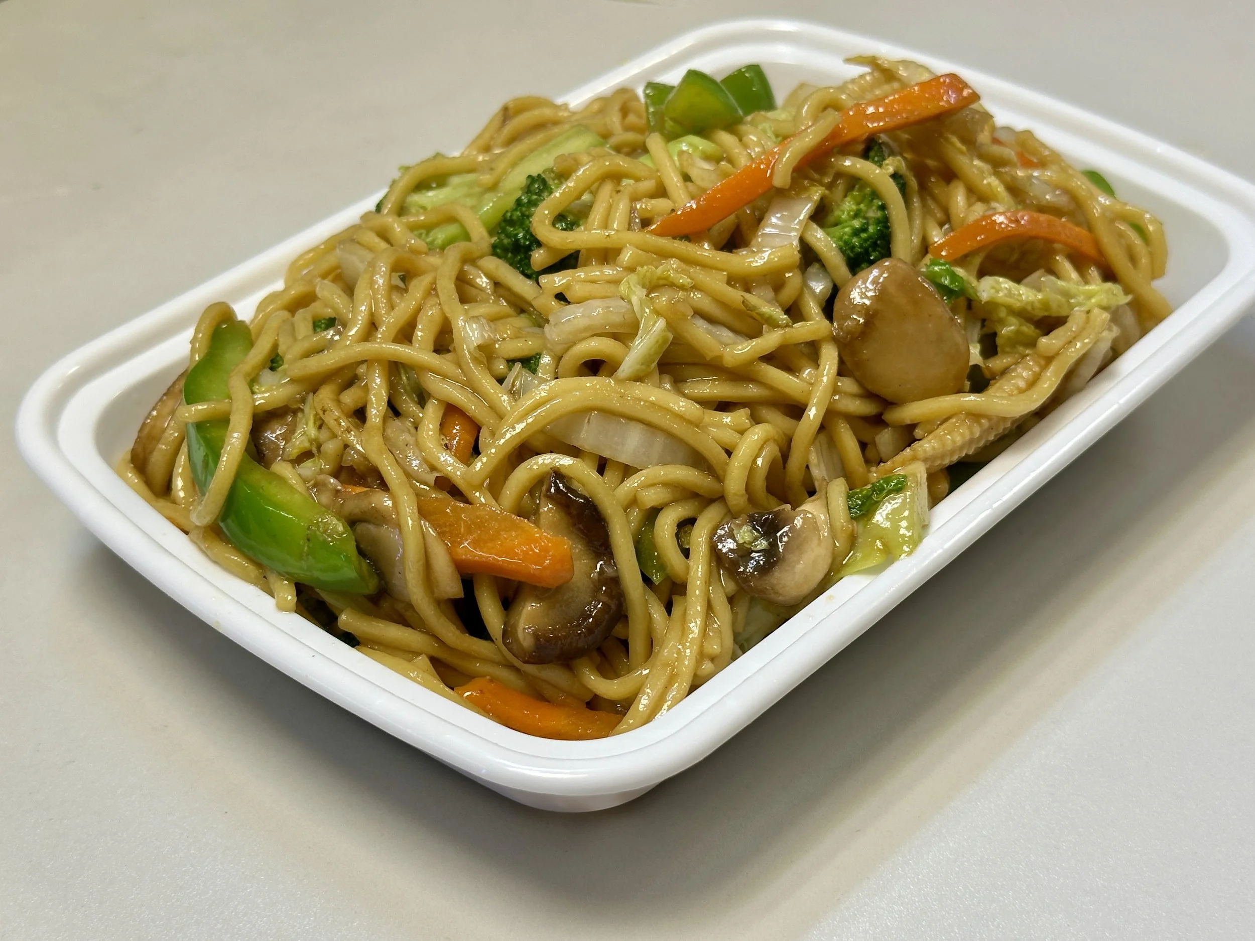 A rectangular white tray filled with stir-fried noodles with vegetables including broccoli, carrots, green bell peppers, and mushrooms.