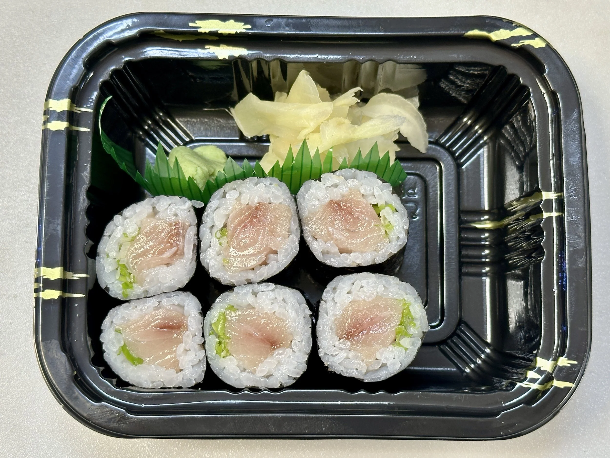 Six sushi rolls with raw fish and rice, garnished with shredded ginger and wasabi in black plastic takeout container