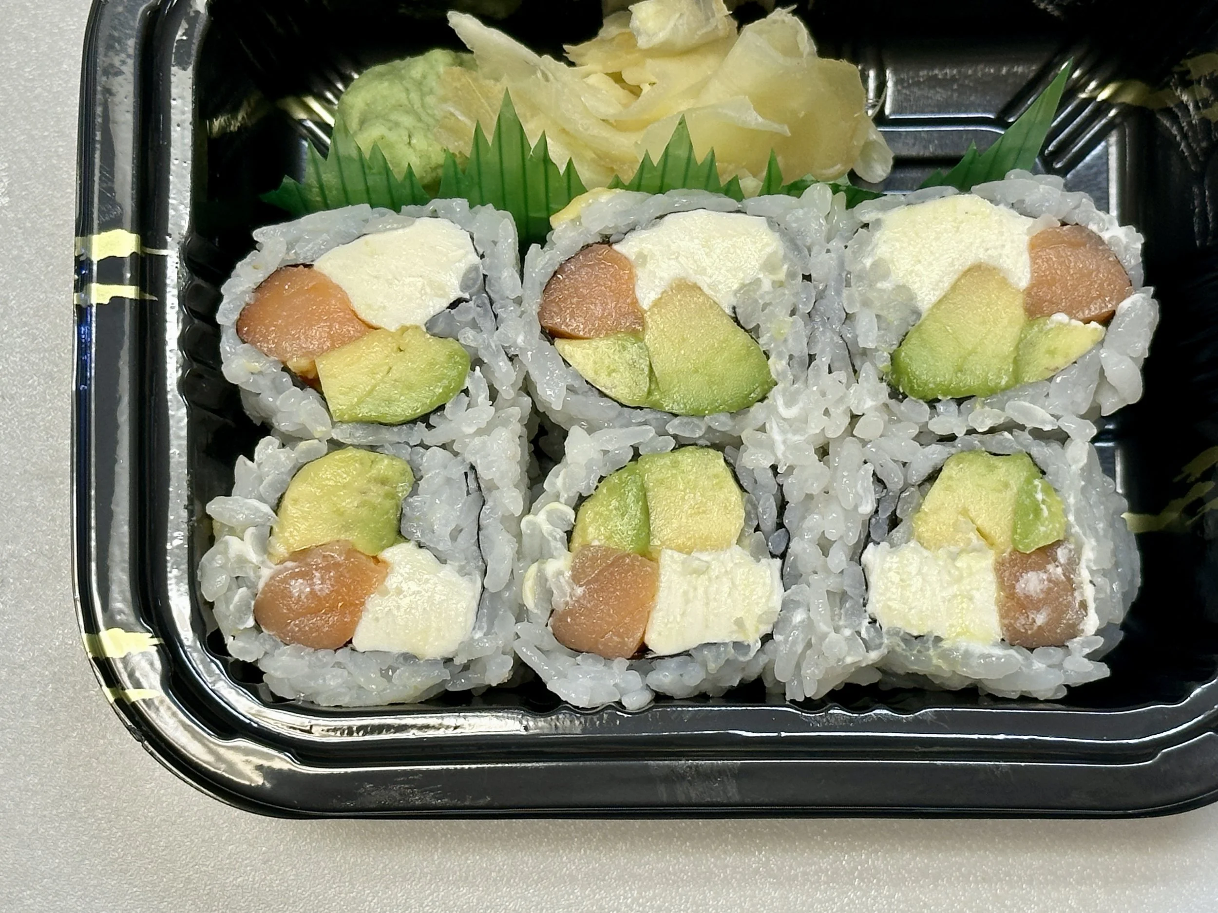A black plastic tray of six sushi rolls filled with avocado, salmon, and cream cheese, garnished with green plastic grass, wasabi, and pickled ginger.
