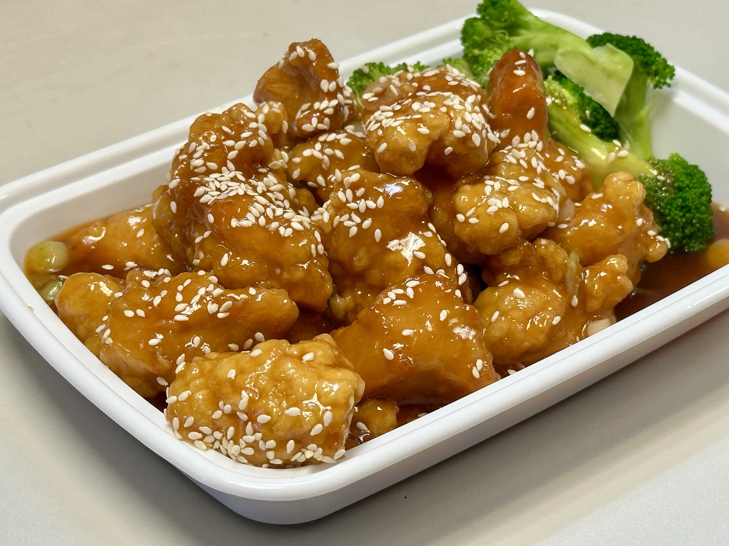 Orange chicken with sesame seeds served with broccoli in a white rectangular dish.
