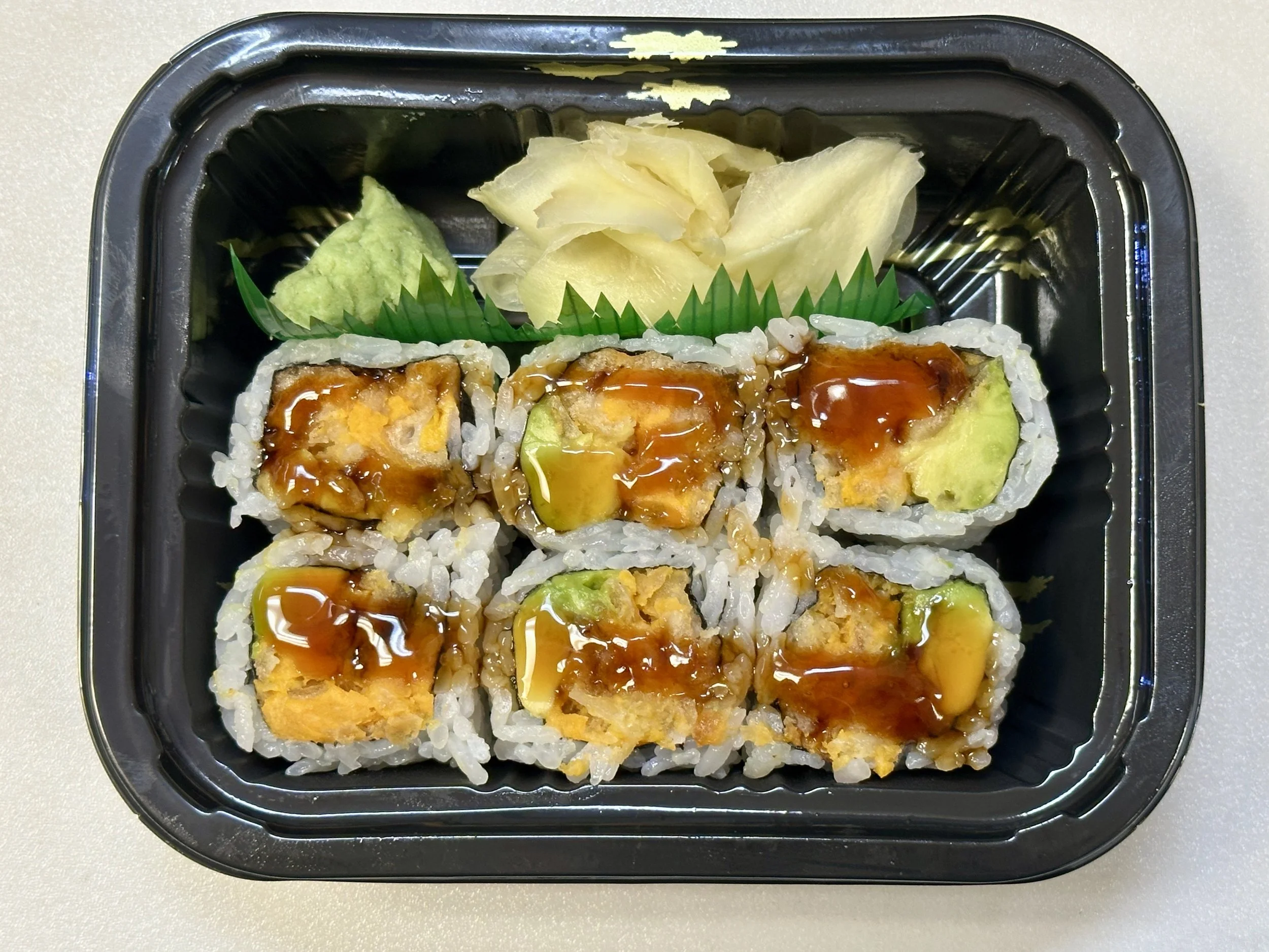 Sushi roll with tempura filling topped with eel sauce, accompanied by pickled ginger and wasabi, in a black plastic takeout container.