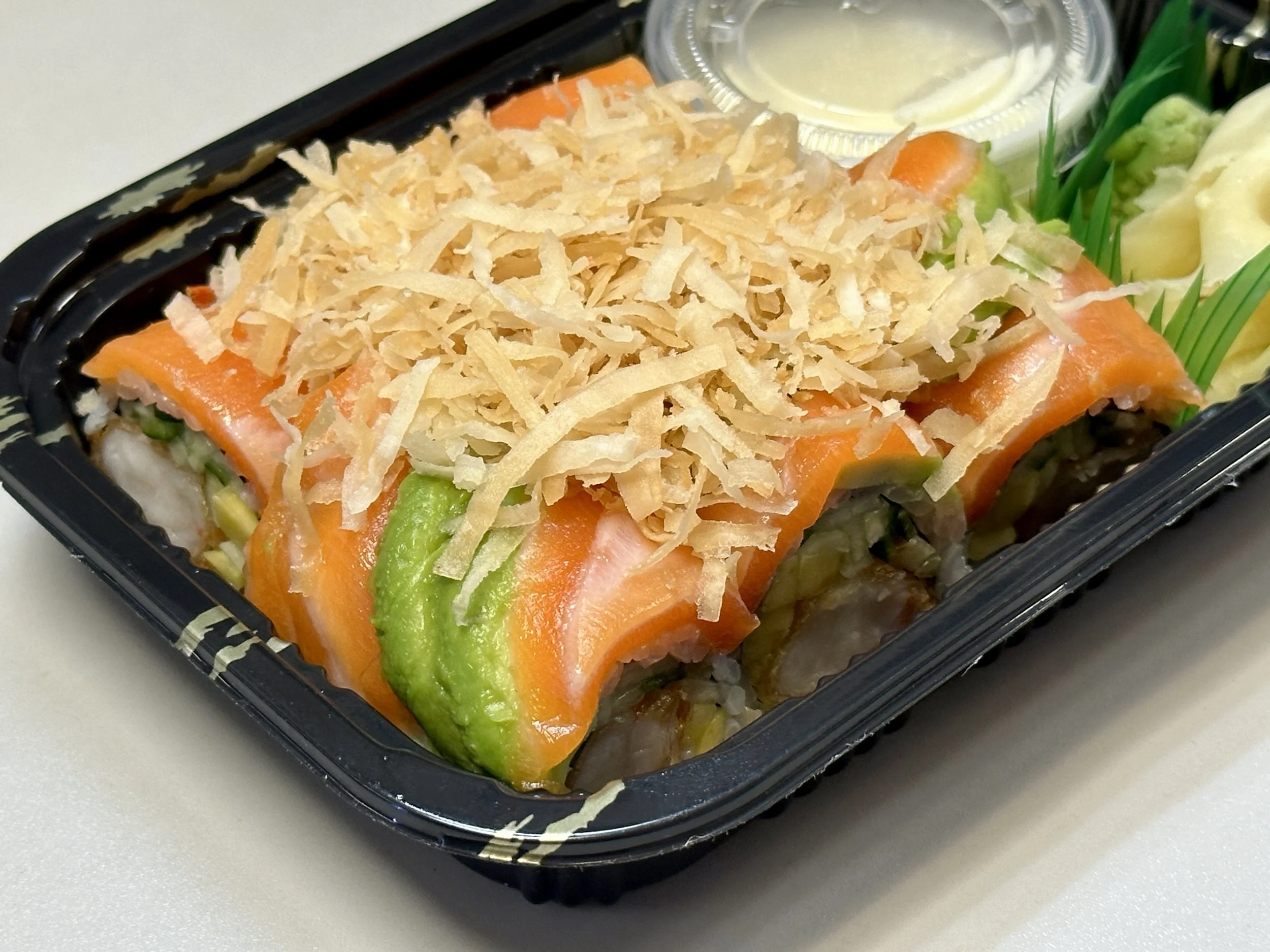 Close-up of a pre-packaged sushi roll topped with shredded cheese, with slices of avocado and salmon visible, along with a small container of dipping sauce.