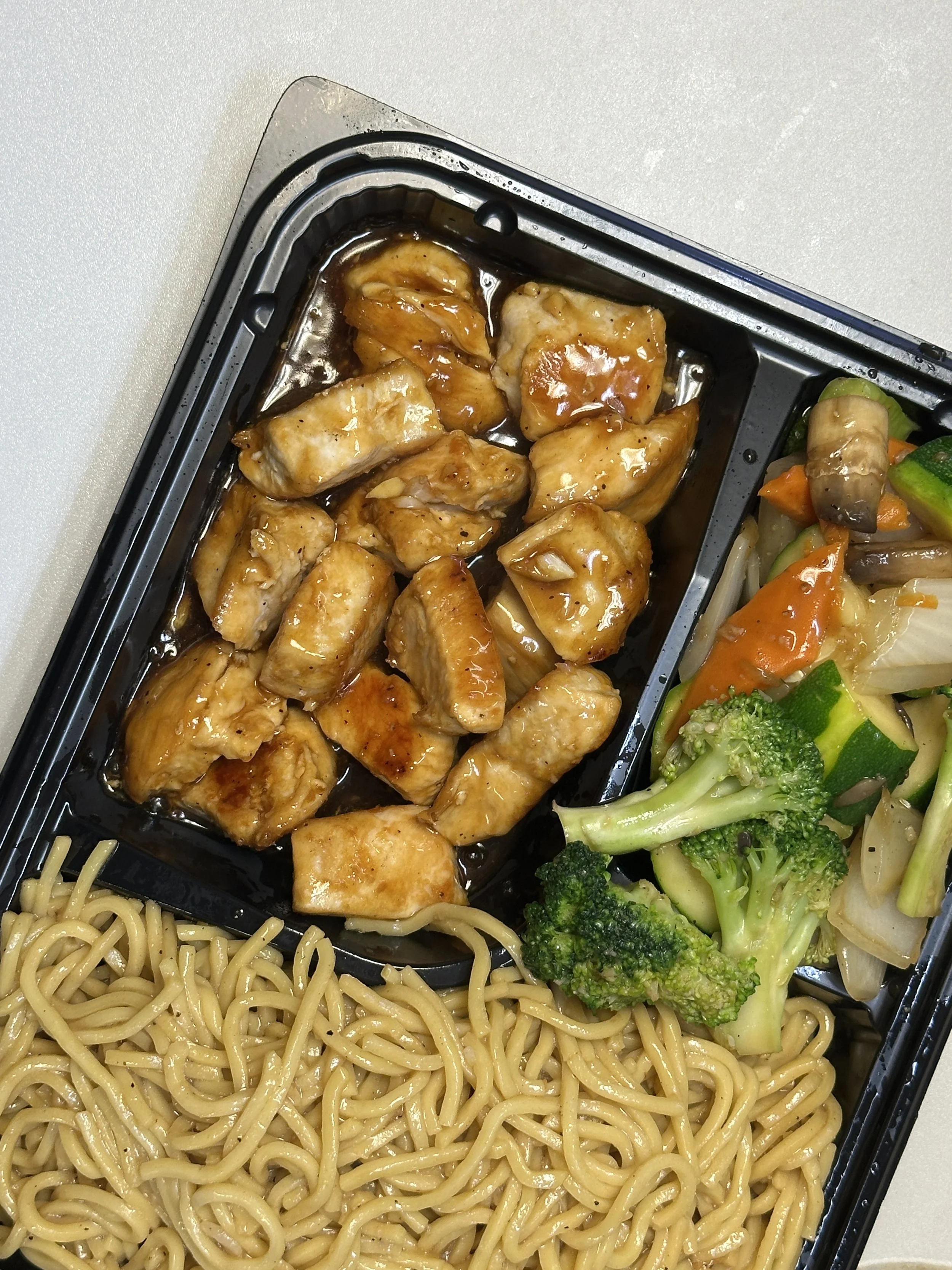 Asian-style meal with stir-fried chicken in teriyaki sauce, cooked broccoli, mixed vegetables, and noodles in a black divided takeout container.