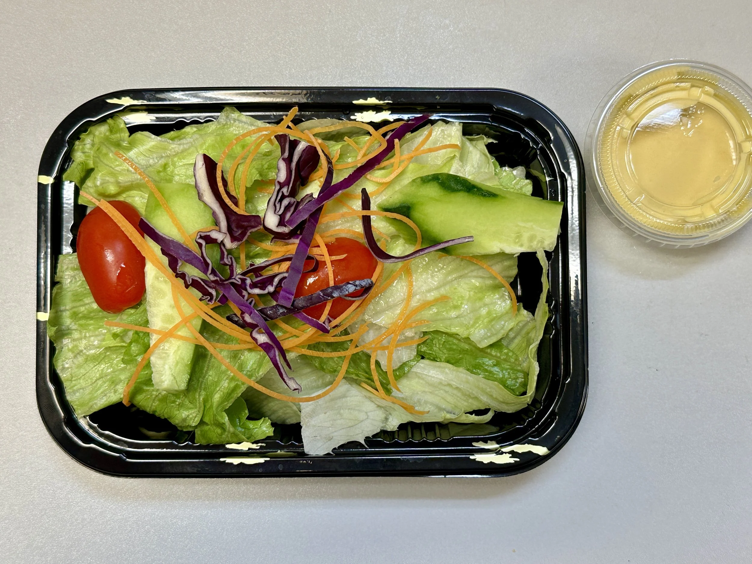 Tray of fresh salad with lettuce, cherry tomatoes, shredded carrots, purple cabbage, cucumber, with a small cup of salad dressing on a light-colored surface.
