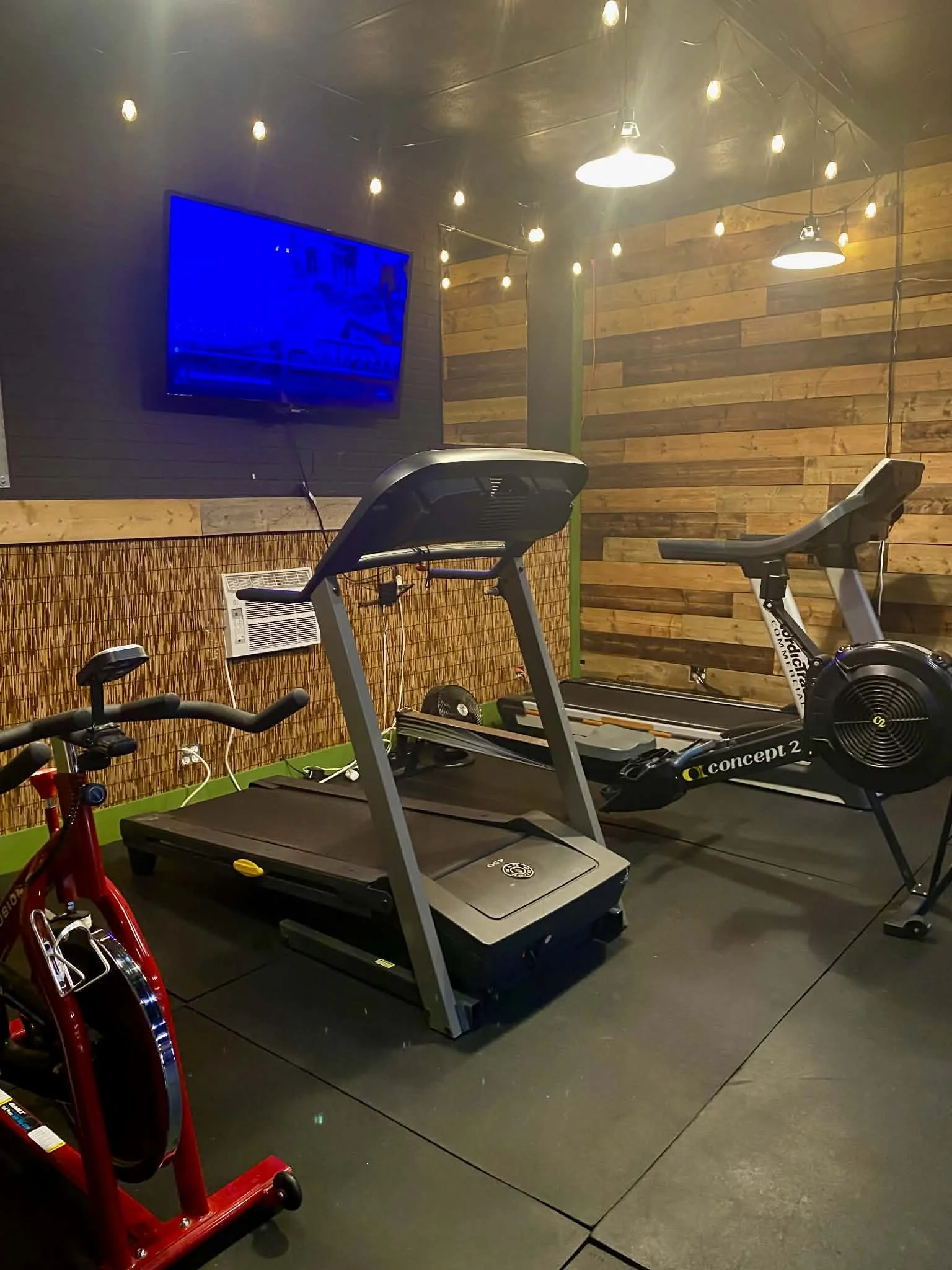 Home gym with treadmill, stationary bike, and television on a dark wall, wooden wall decor, and string lights overhead.