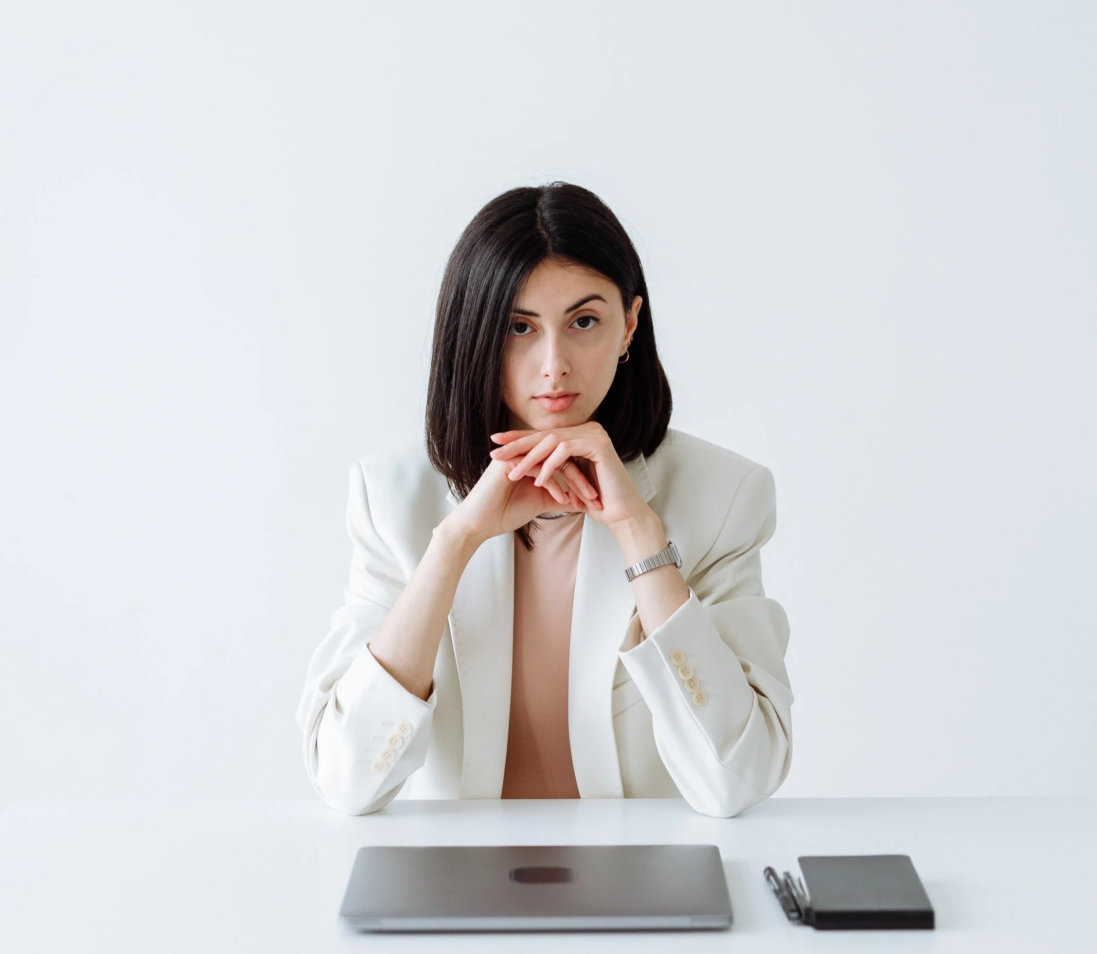 A woman with dark hair wearing a white blazer sitting at a white desk with a laptop, pen, and notebook against a white background.