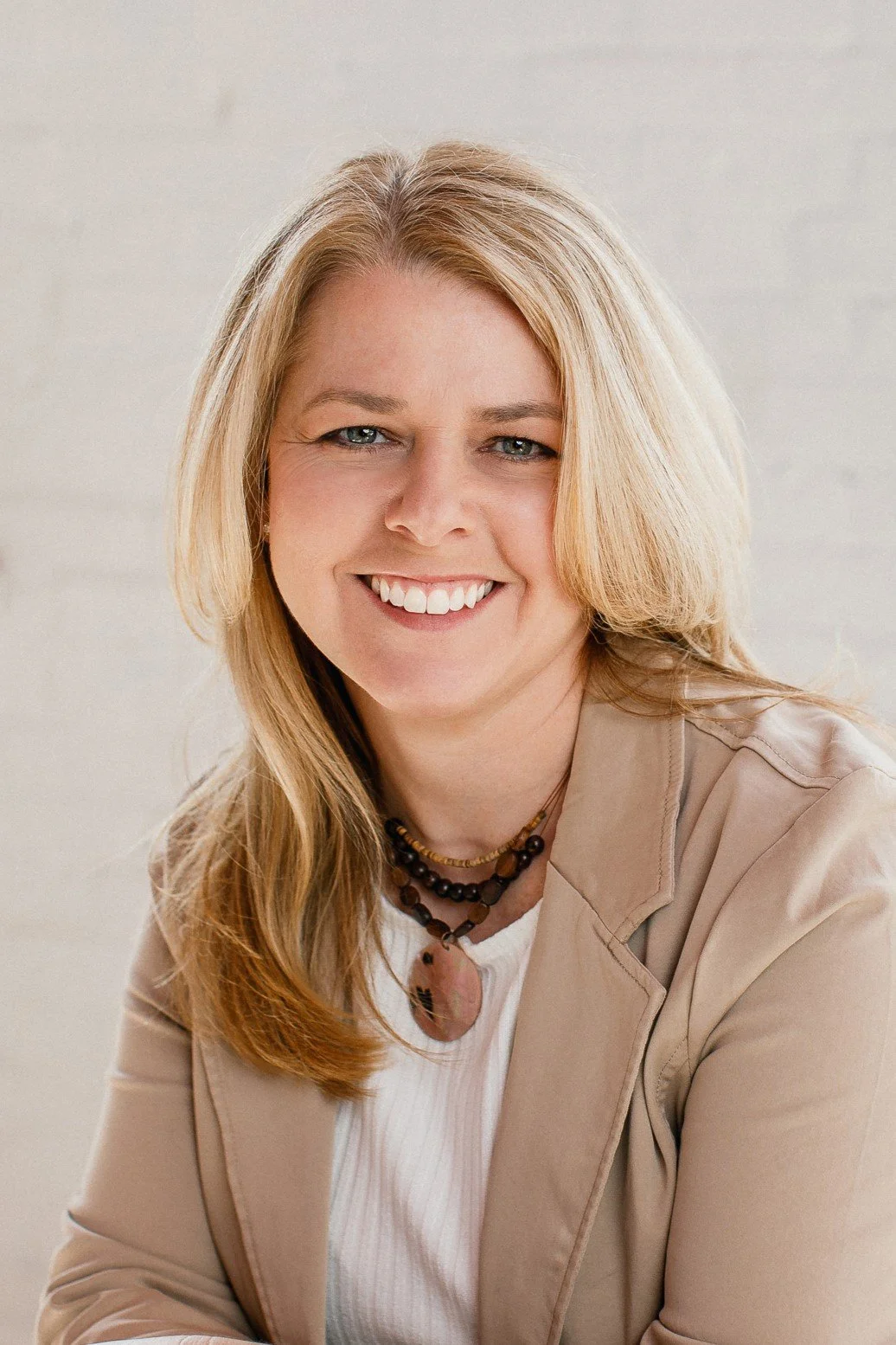 A woman with blonde hair smiling, wearing a beige jacket, a white top, and beaded necklaces, sitting against a light-colored background.