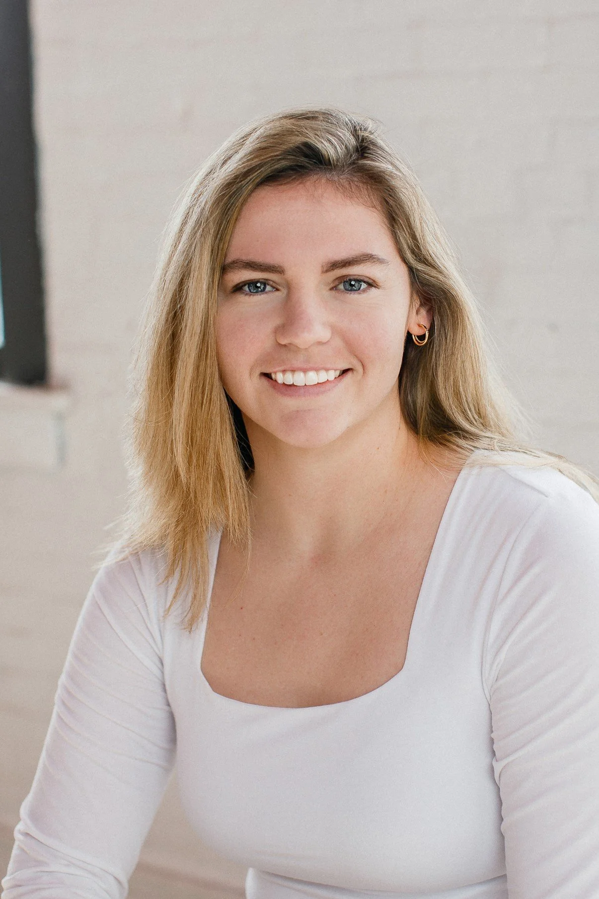 A young woman with blonde hair, wearing a white top, smiling at the camera with blue eyes and gold hoop earrings, indoors with a light-colored background.