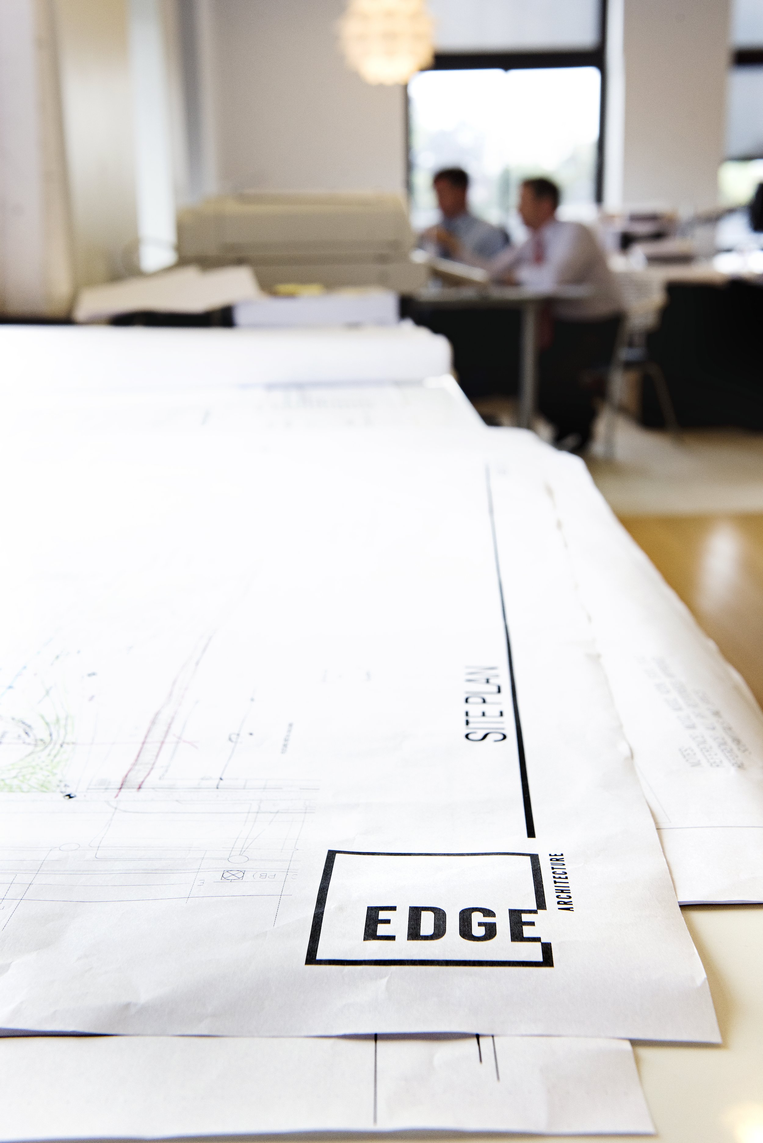 Architectural blueprints on a table in a modern office. Two men are working at a desk in the background, with large windows and natural light.