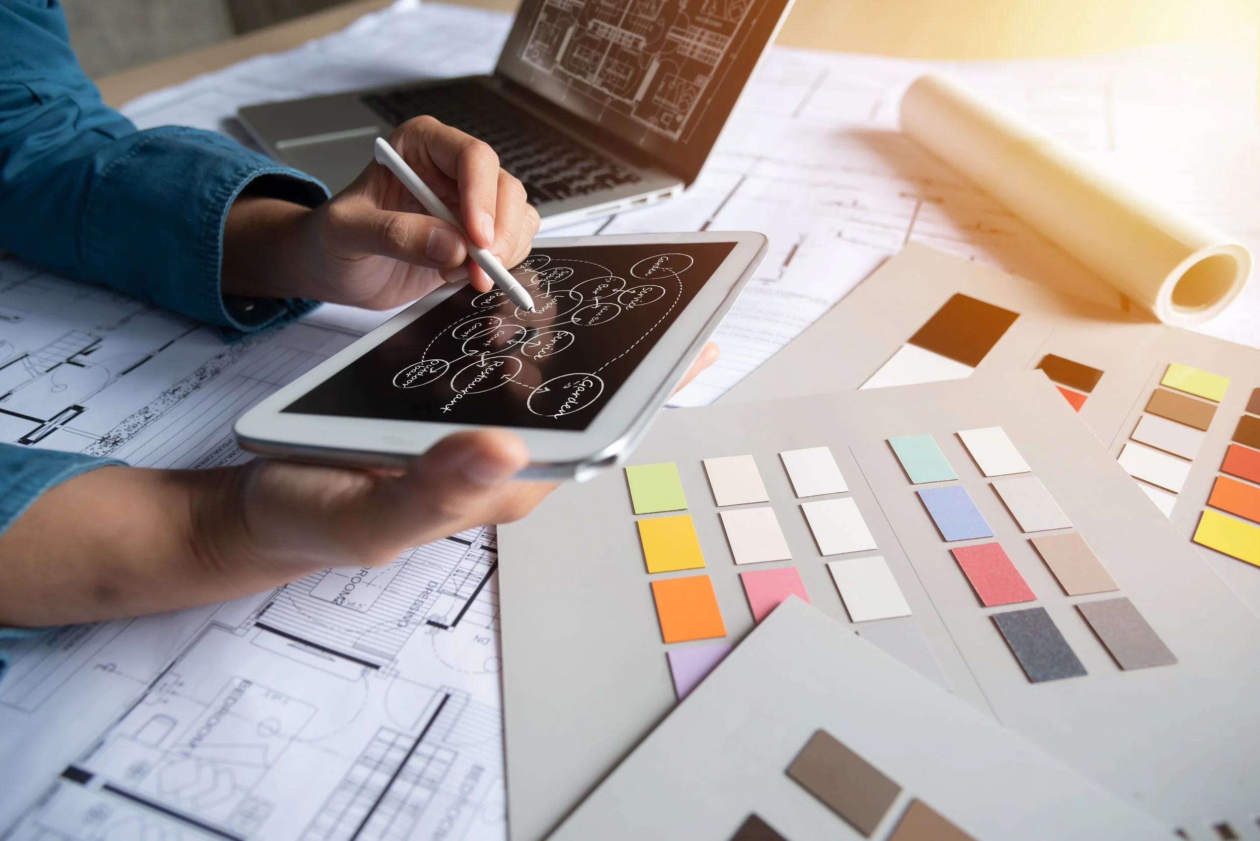 A person using a stylus on a digital tablet with architectural drawings and color samples on a table, with a laptop in the background.