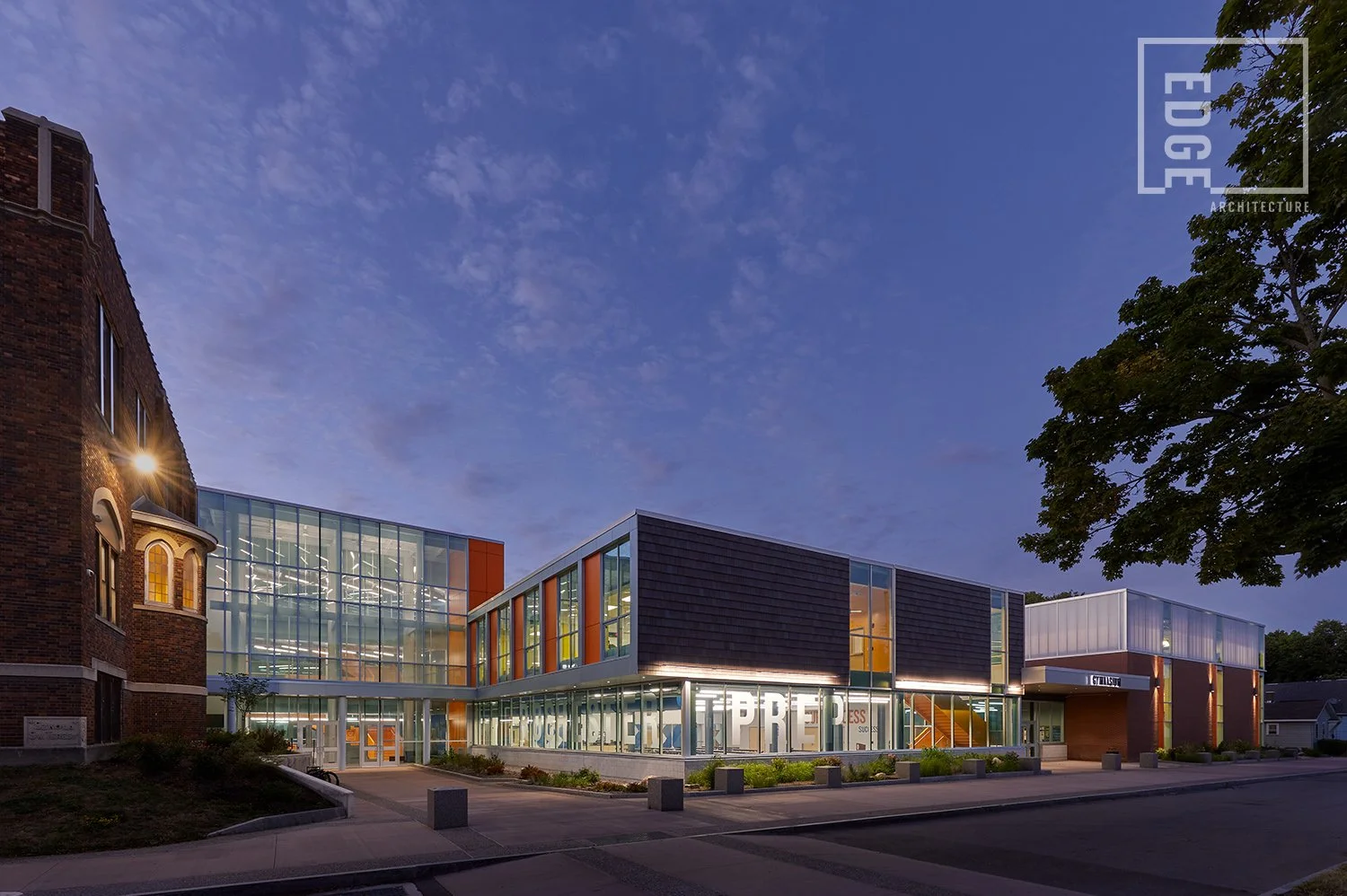 A modern glass and metal building with large windows and colorful exterior accents, illuminated at dusk, neighboring an older brick building, with trees and a sidewalk in front.
