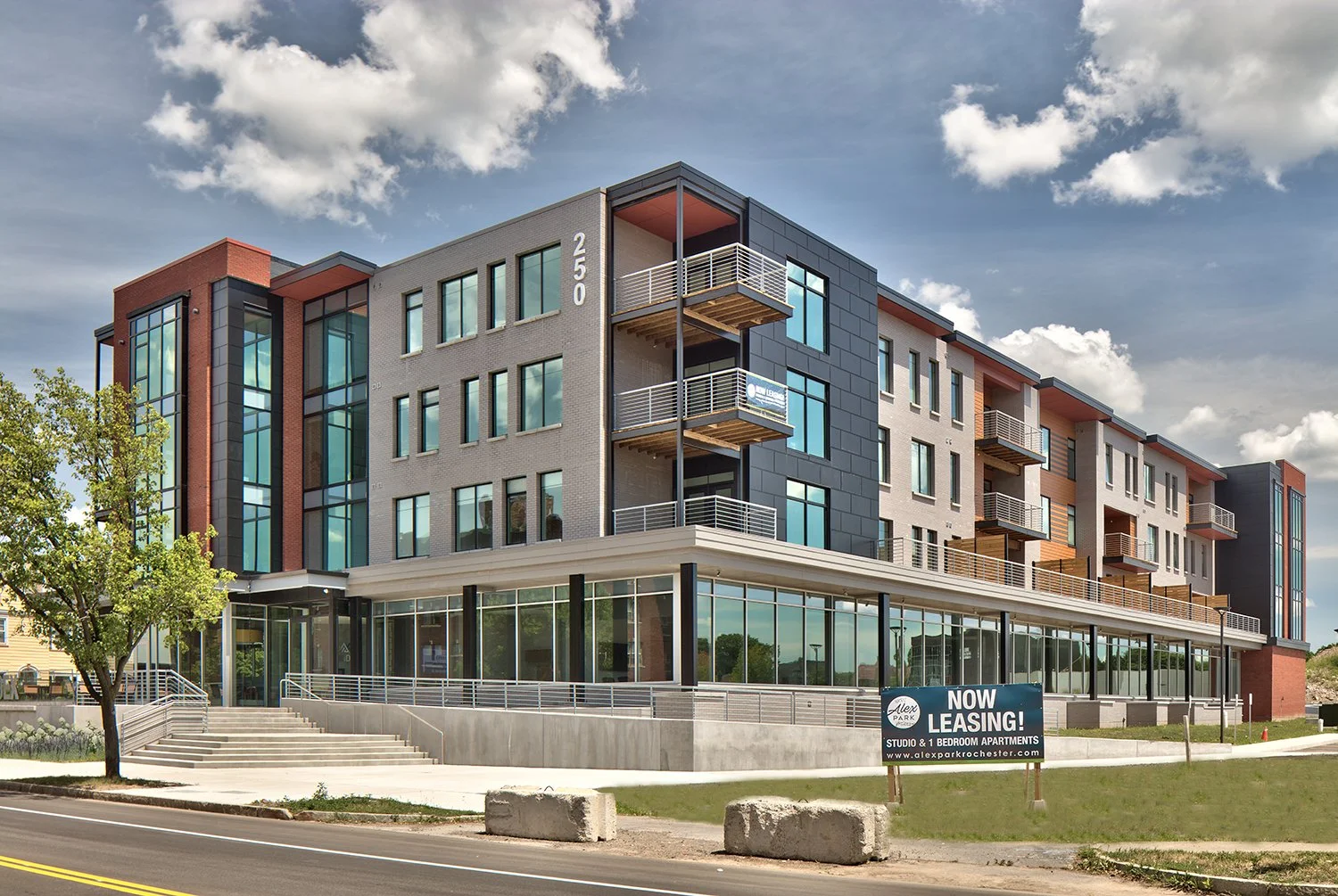Modern multi-story apartment building with multiple balconies, large glass windows, and a sign indicating now leasing for studio and one-bedroom apartments.
