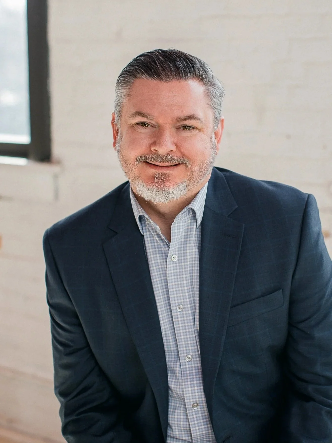 A middle-aged man with gray hair and beard, wearing a navy blazer and light blue checked shirt, sitting indoors near a window.