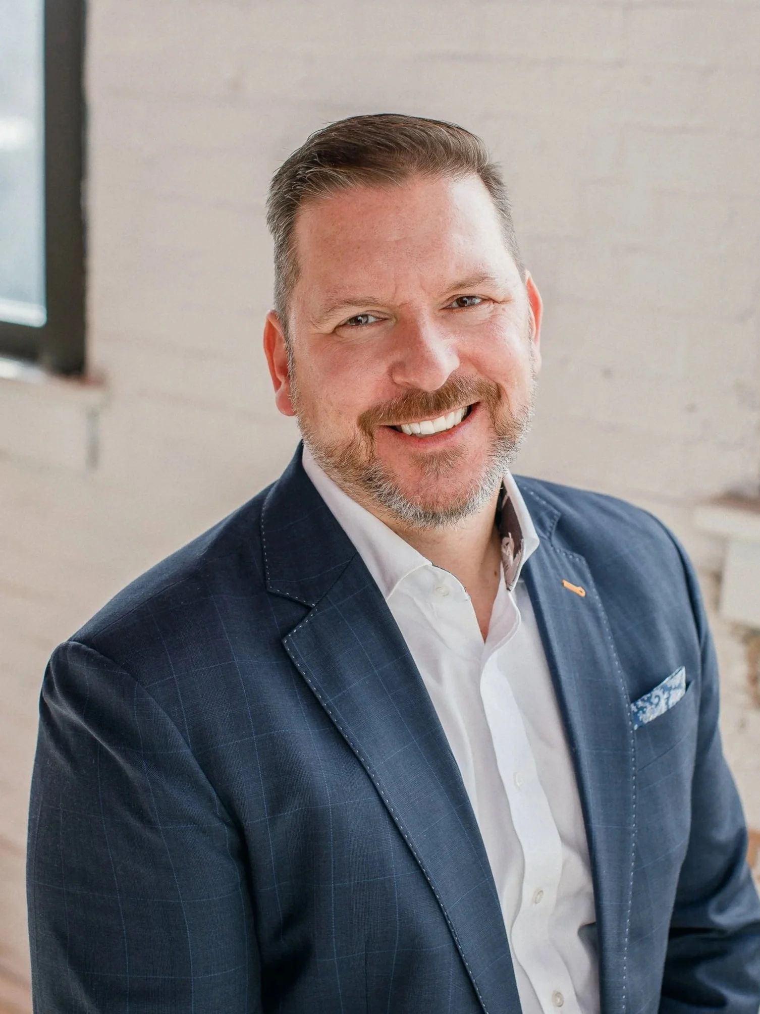 A middle-aged man with brown hair and a beard, wearing a dark blue suit and white shirt, smiling at the camera in an indoor setting.