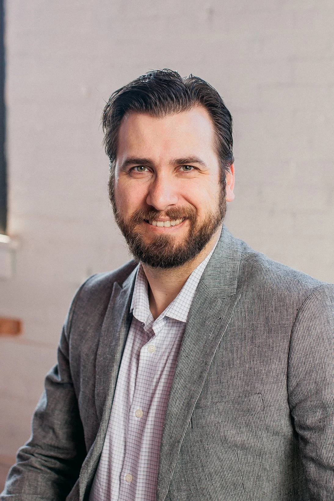 A smiling man with dark hair and a beard, wearing a gray suit jacket and a white checkered shirt, in an indoor setting with a blurred background.