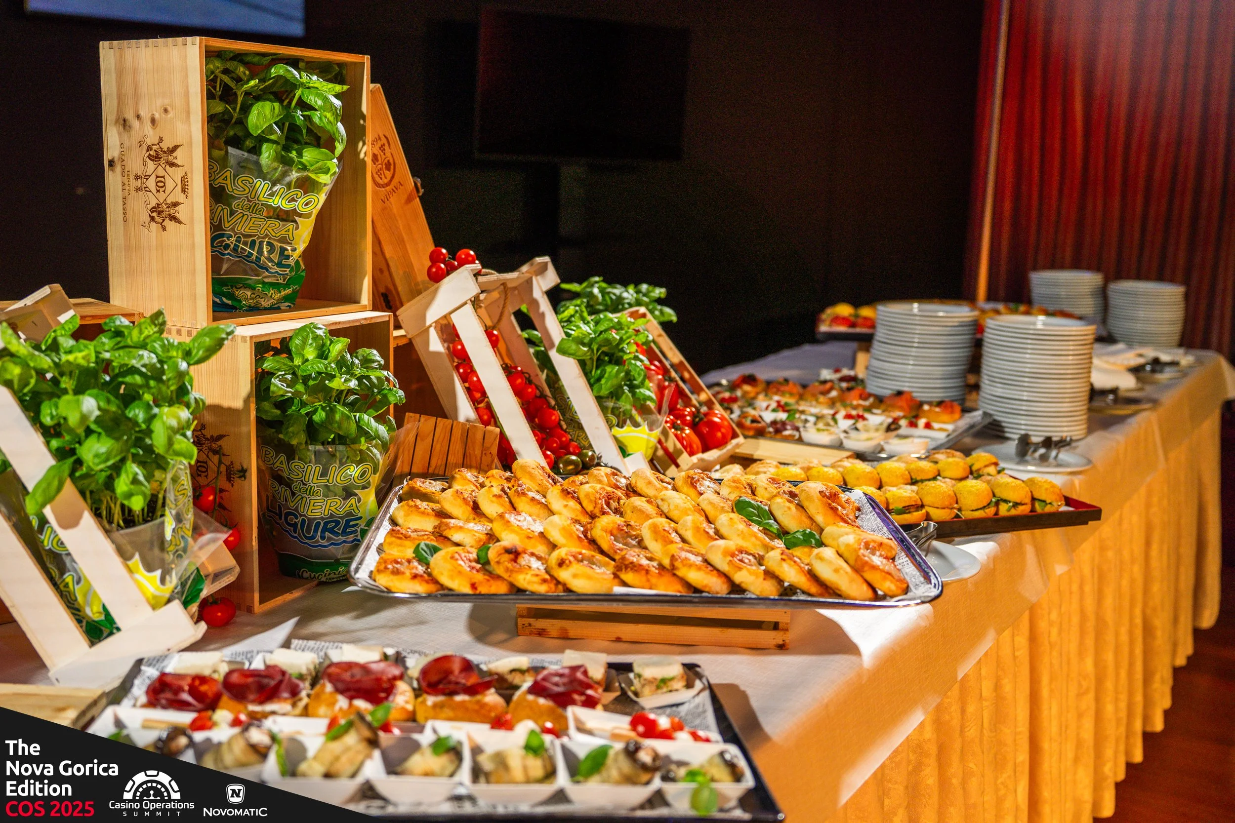 Buffet table with assorted appetizers, finger foods, and desserts, including baked items, vegetables, and small plates, arranged on trays and plates, with stacks of white plates and decorative crates of basil and cherry tomatoes in the background.