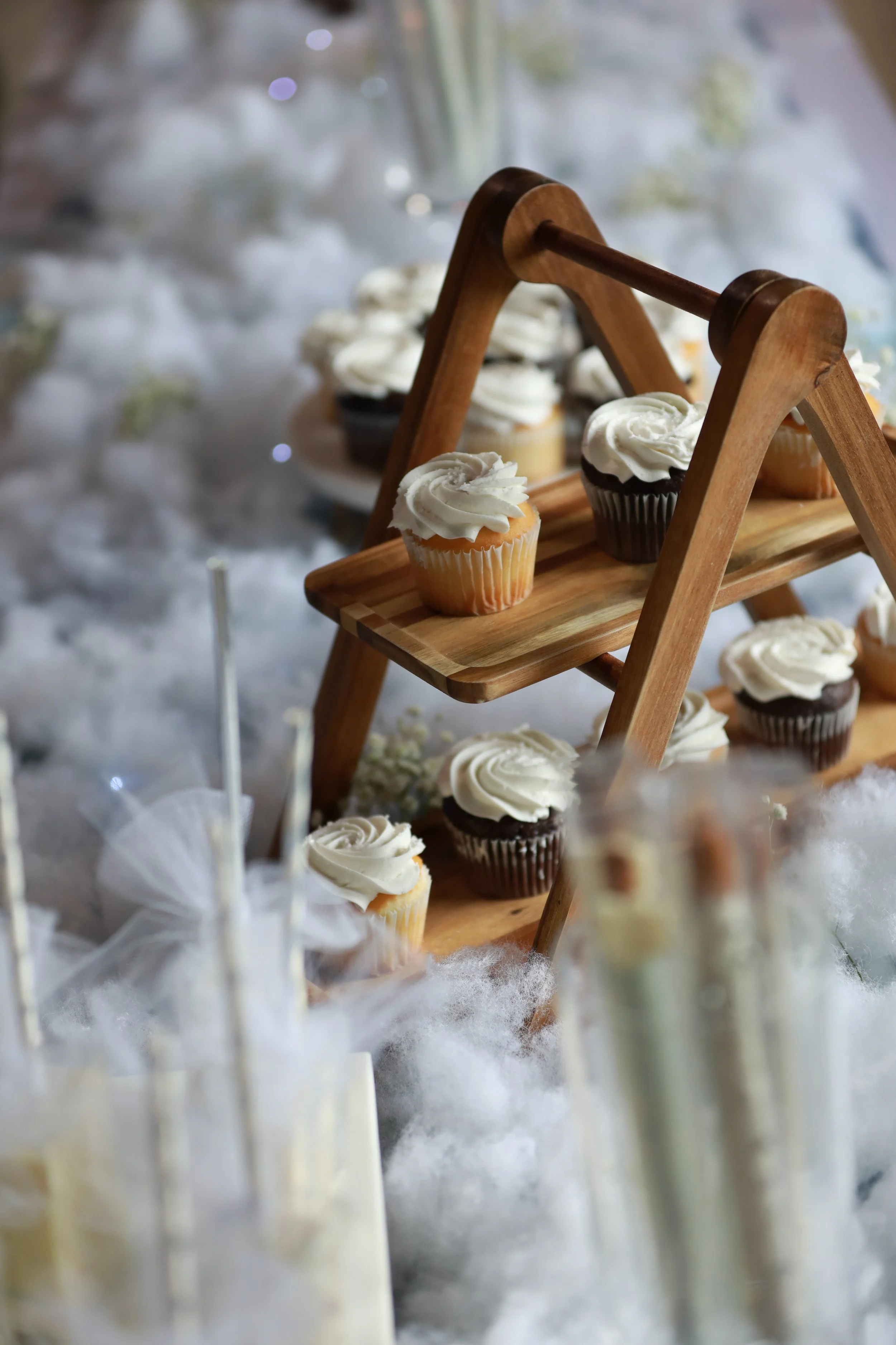 Cupcakes with white and chocolate frosting on a wooden display stand, surrounded by cotton-like decorations, with some holiday-themed treats in the background.
