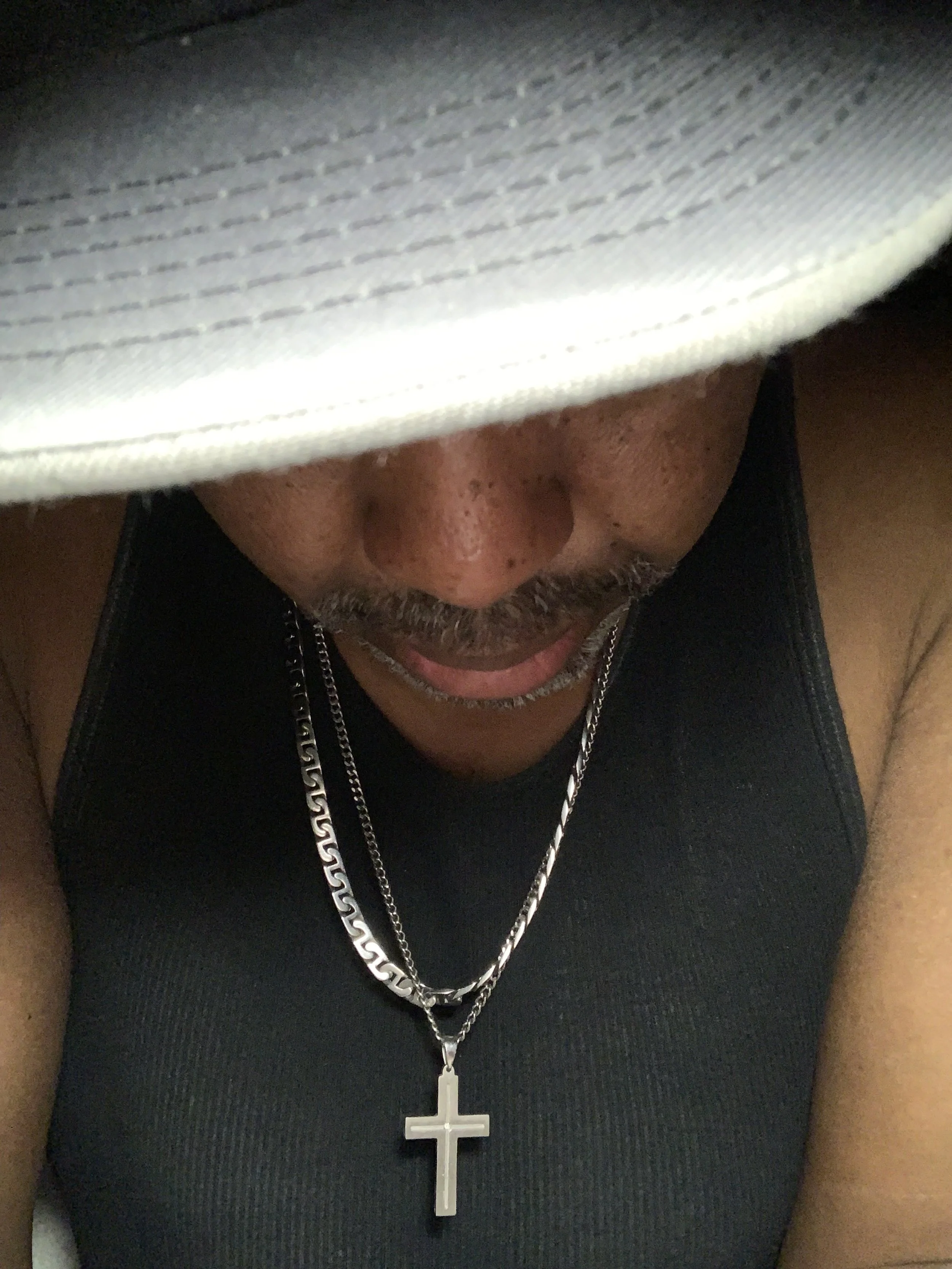 A close-up of a man's face partially obscured by a white baseball cap, wearing a black sleeveless shirt and silver chains with a cross pendant.