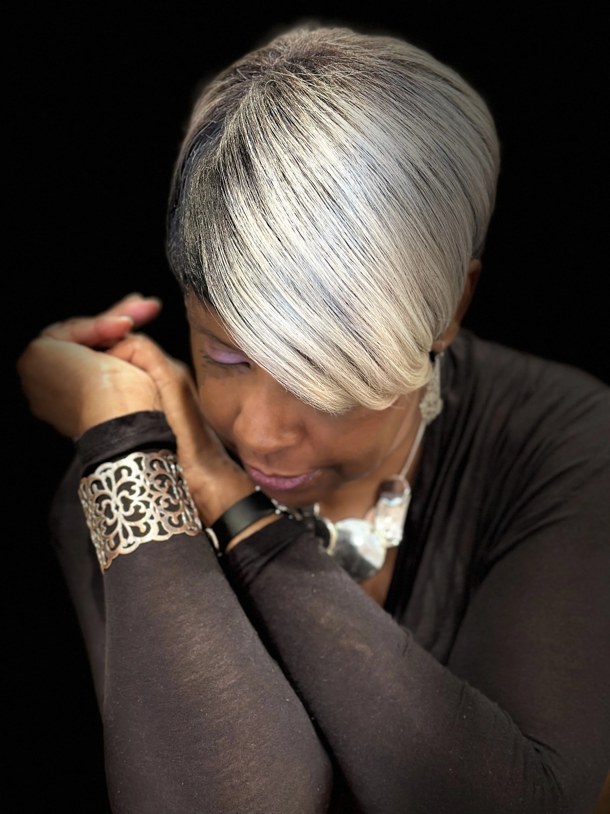 Woman with short, platinum blonde hair, wearing black top, jewelry, and braces, resting her head on her crossed arms against a dark background.