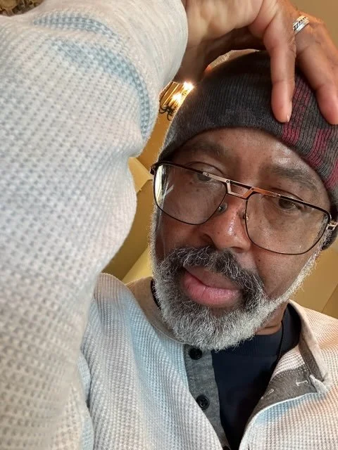 A man with glasses and a gray beard wearing a beanie and a white sweater taking a selfie indoors.