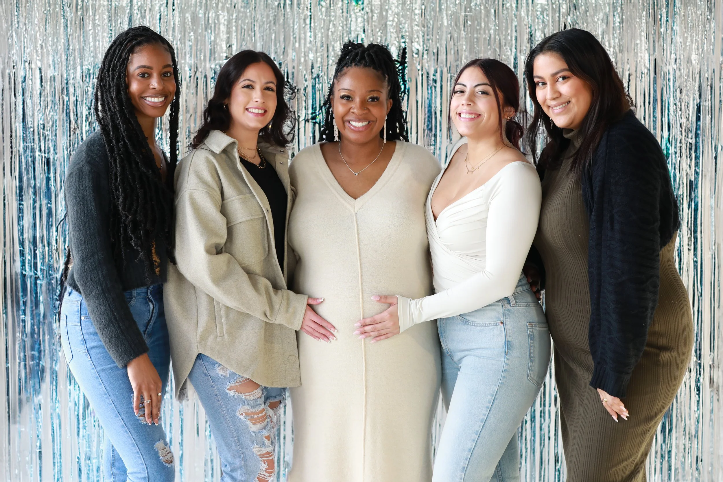 Group of six women standing together in front of a silver metallic backdrop, smiling and touching her pregnant belly, dressed casually.