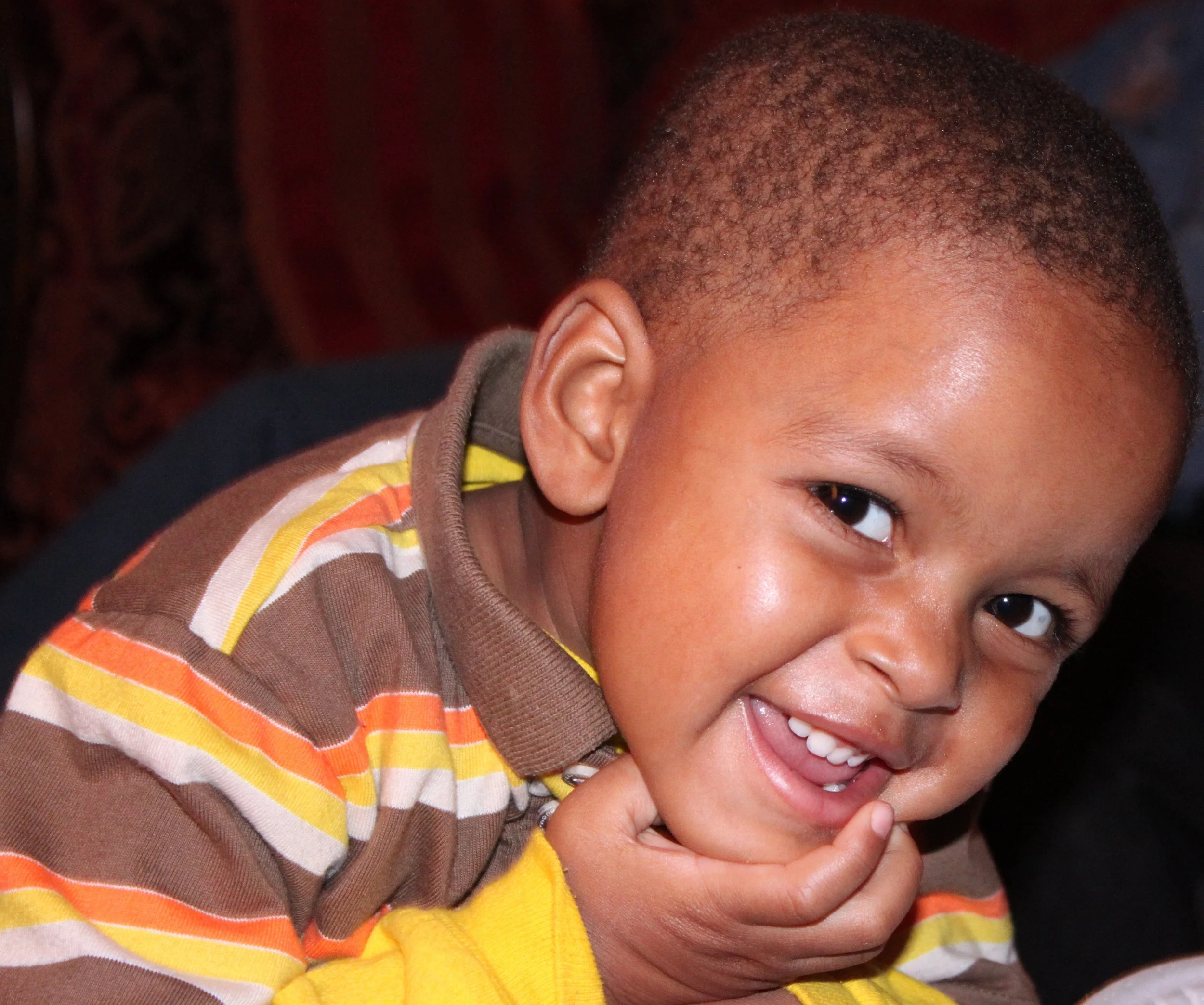A young boy with short hair and medium skin tone smiling and holding his chin with his hand, wearing a brown and yellow striped shirt.