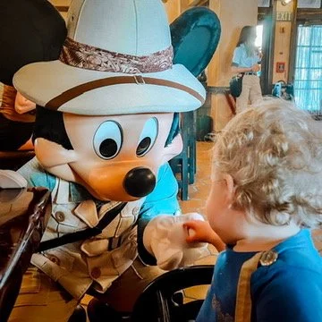 Mickey Mouse dressed as an explorer talking to a young boy in a restaurant.