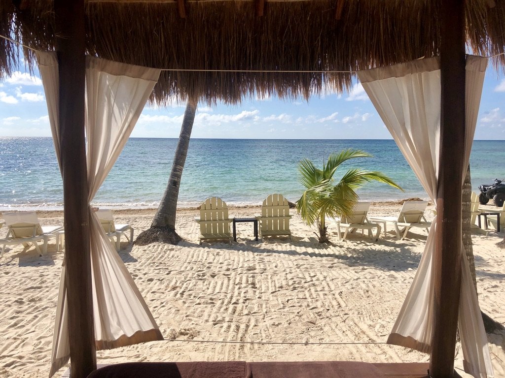 Beach scene viewed from under a thatched cabana, with white curtains, a sandy shore, a leaning palm tree, lounge chairs, small palm, ocean, and blue sky with clouds.