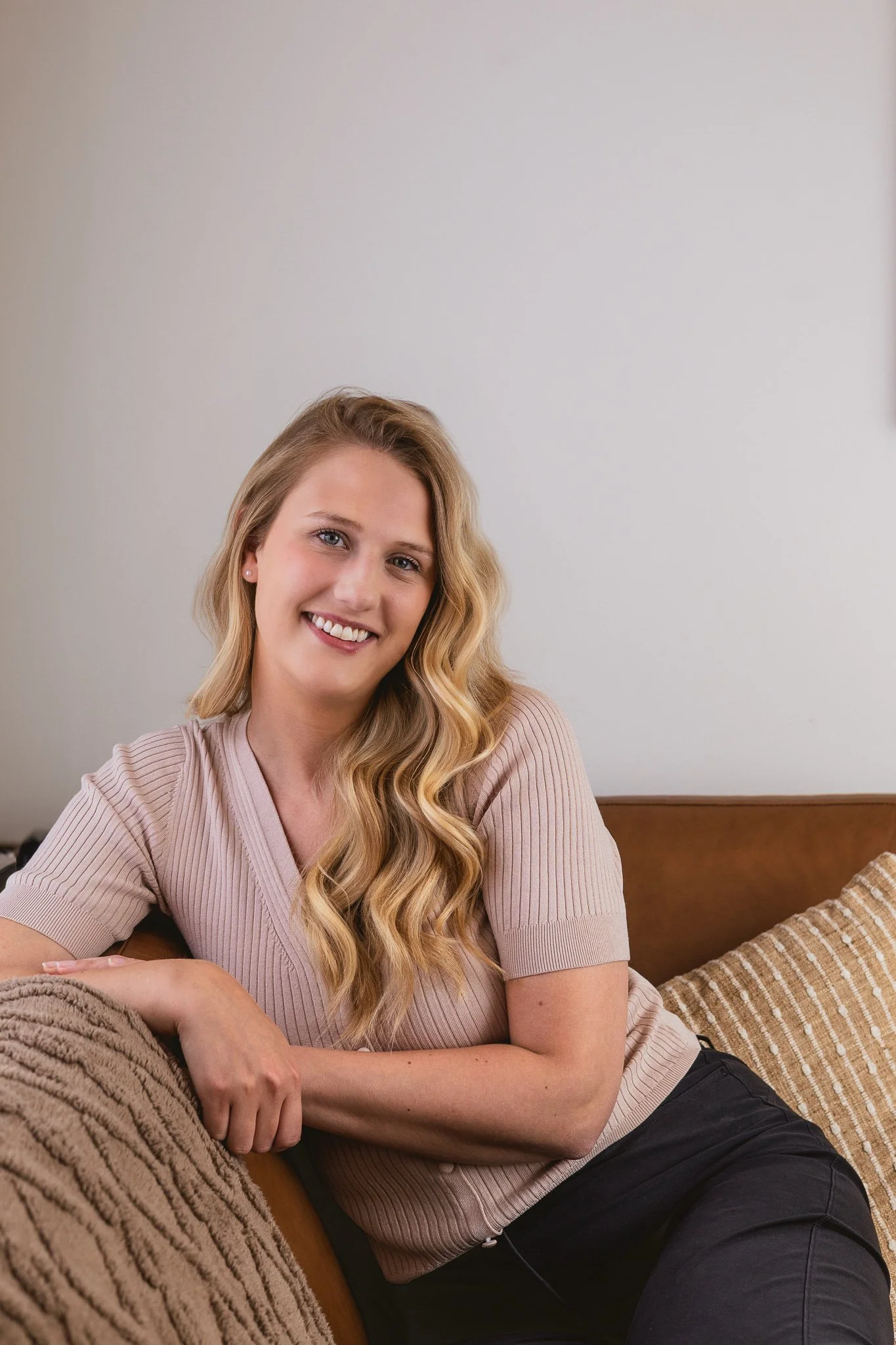 A woman with long wavy blonde hair smiling, sitting on a brown sofa with a beige blanket and a striped pillow.
