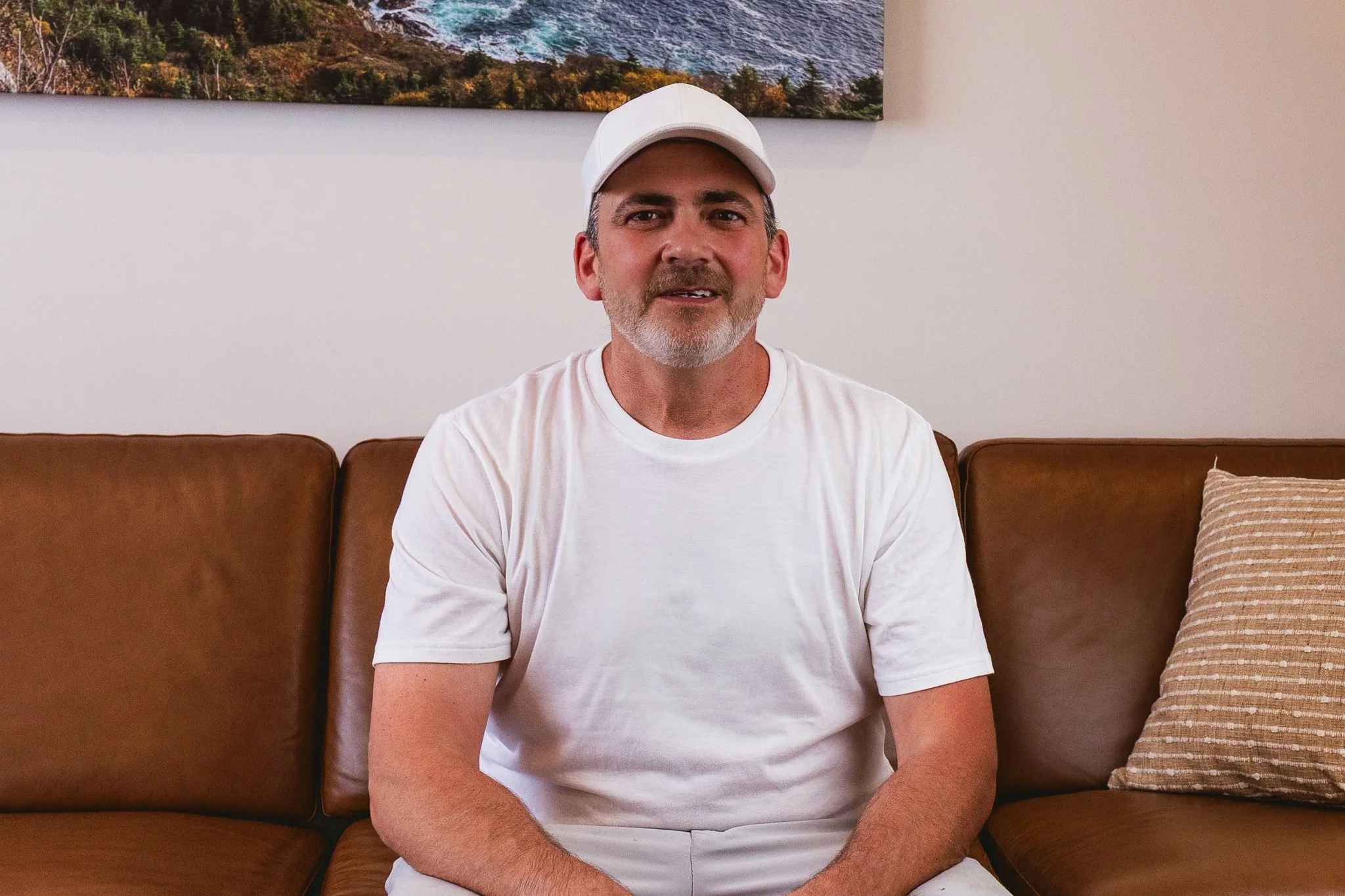 A middle-aged man with a beard and mustache wearing a white cap and white shirt, sitting on a brown sofa in front of a light-colored wall with a landscape painting above.
