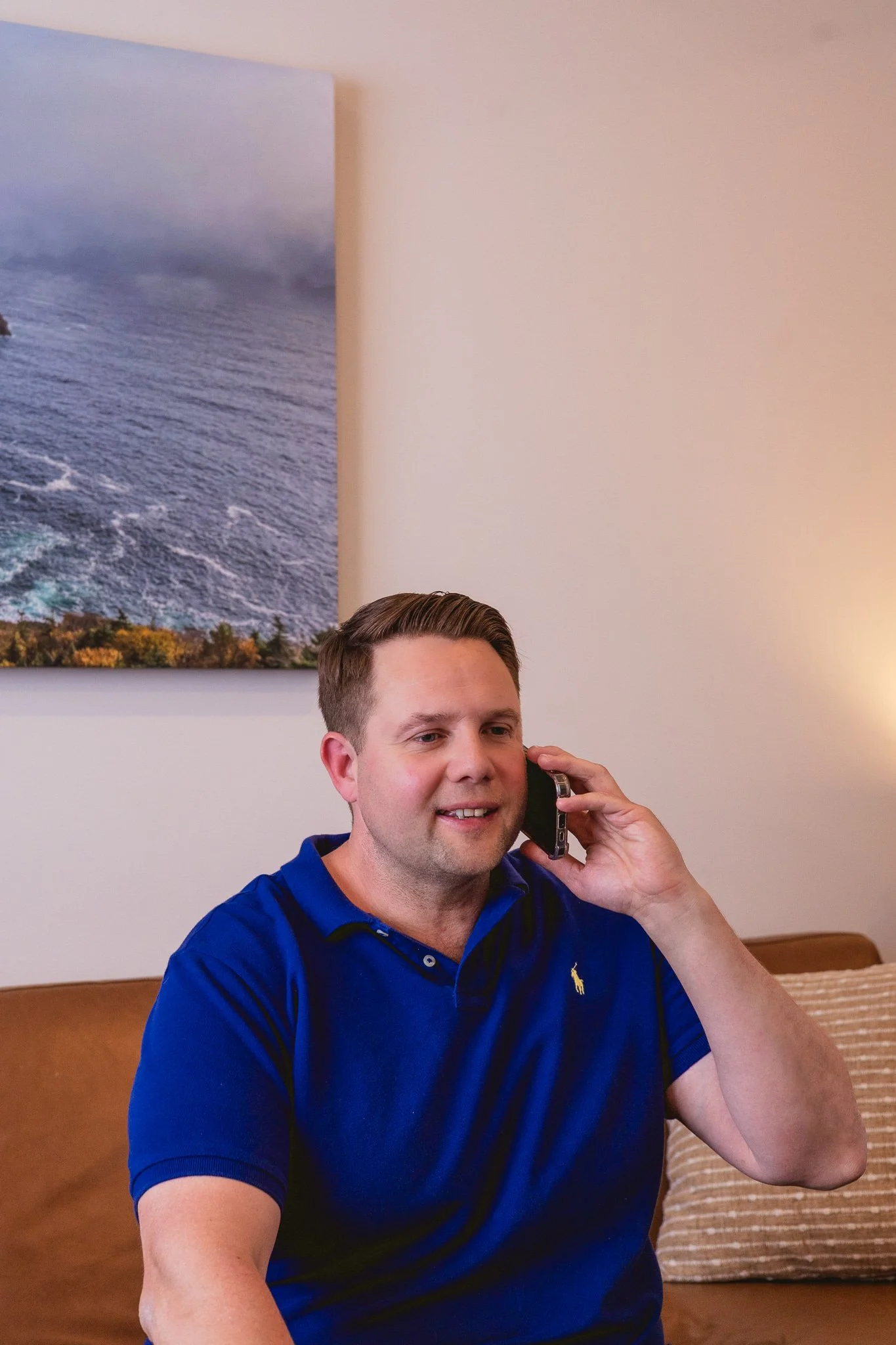 A man with short brown hair and a blue polo shirt talking on a mobile phone while sitting on a beige couch with striped pillows in a living room.