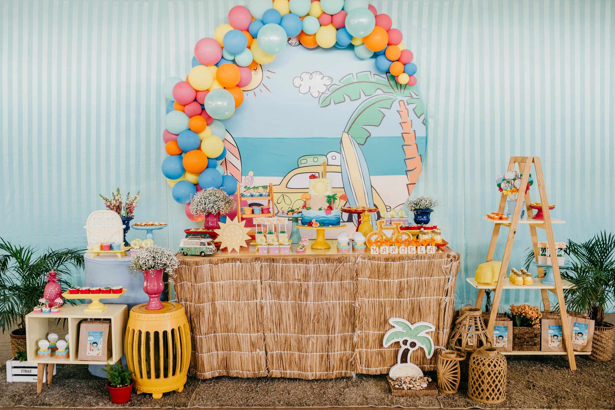 Tropical-themed birthday party table with colorful balloons, desserts, and decorations featuring palm trees, surfboards, and a beach scene backdrop, decorated in summer colors.