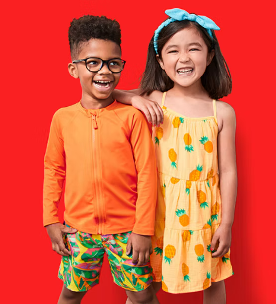 A boy and girl are smiling and laughing together, standing against a red background. The boy wears glasses, an orange jacket, and colorful shorts. The girl wears a yellow pineapple-print dress with a blue bow headband.