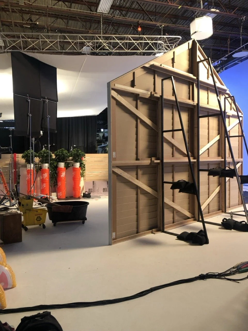 The image shows a partially constructed set or backdrop with a wooden frame and ladder, in a studio or warehouse. There are some potted plants, equipment, and some lighting or support structures in the background.