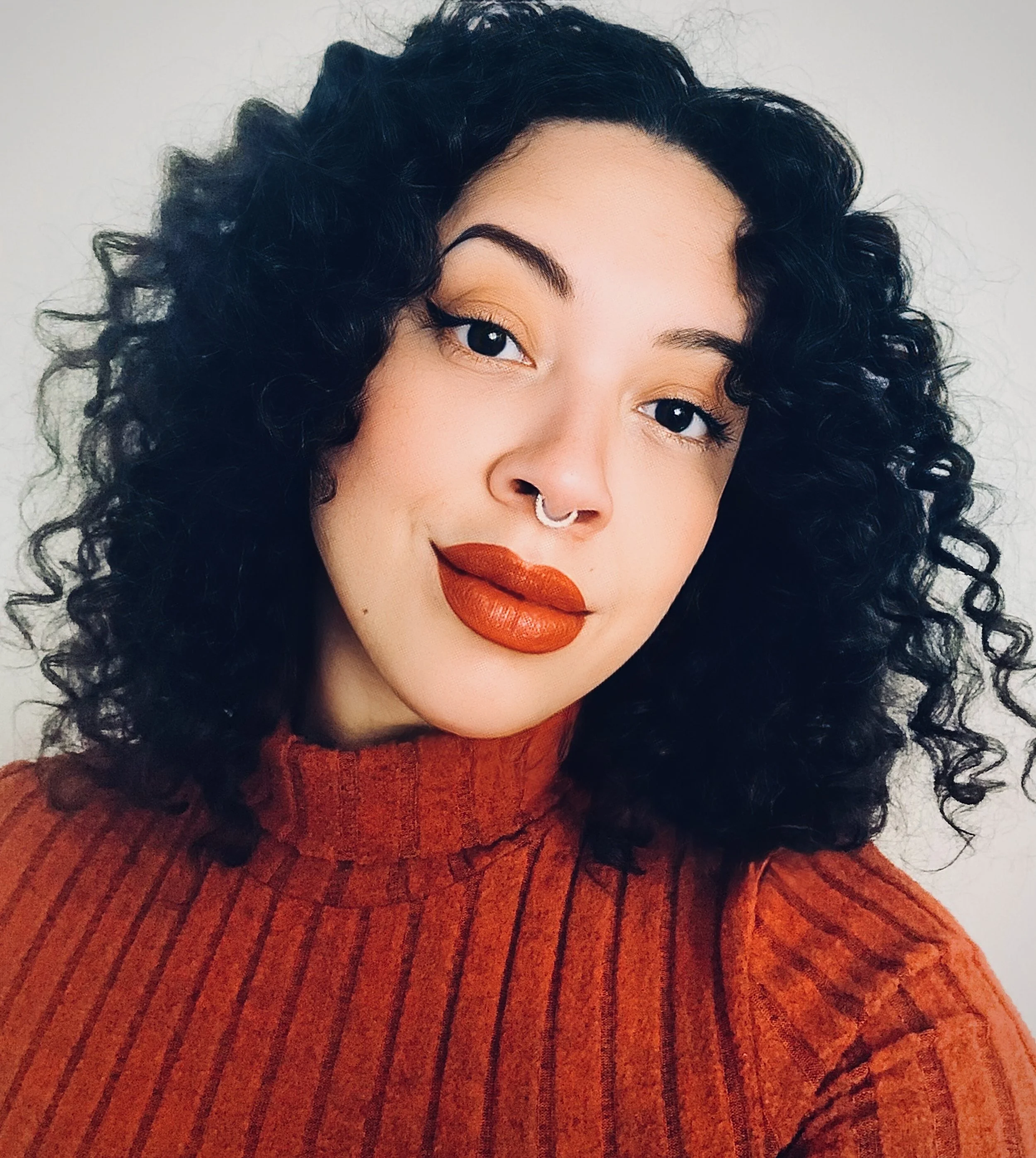 A woman with curly black hair, orange lipstick, and a septum piercing wearing a red ribbed turtleneck sweater.