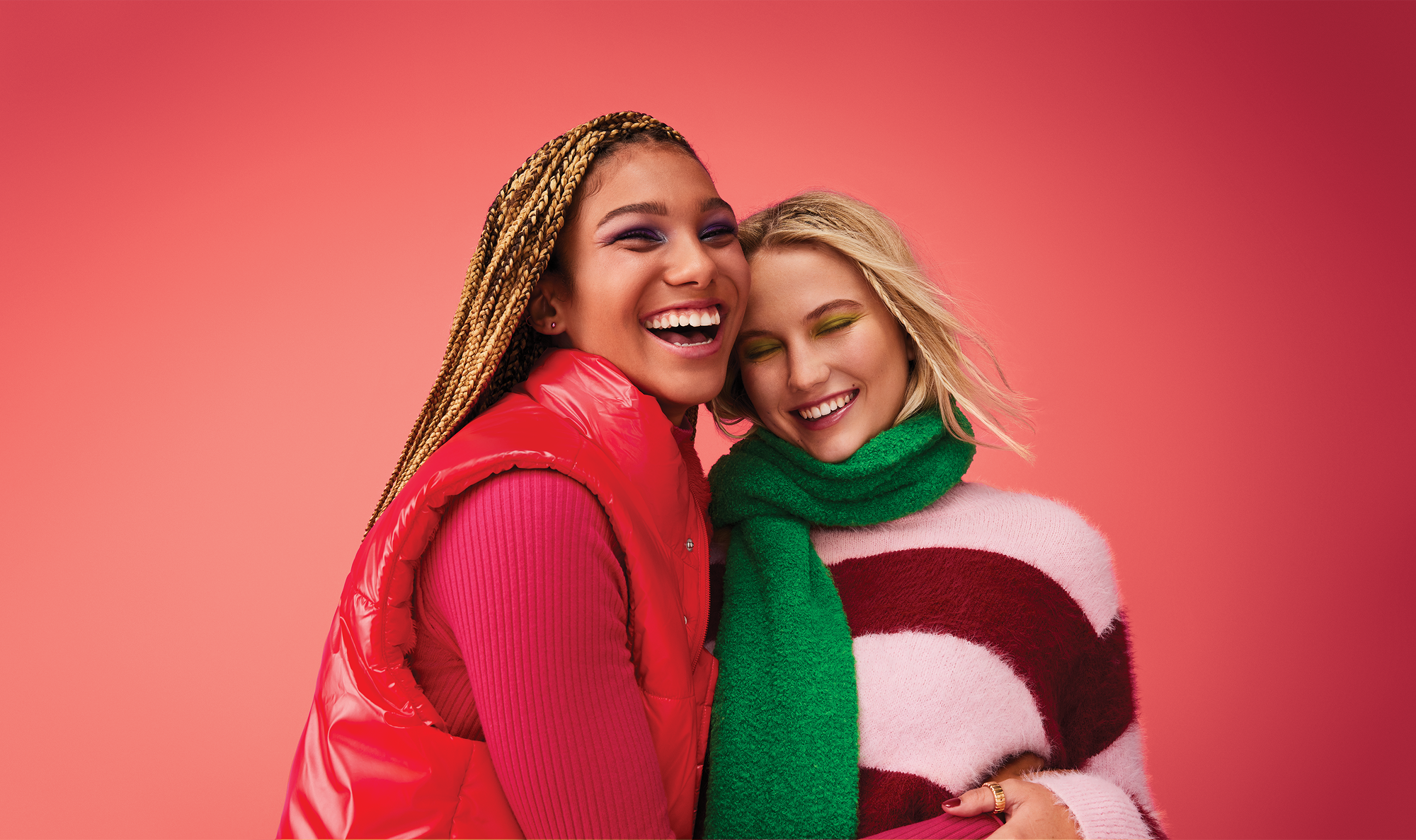 Two women smiling and hugging against a pink background, one wearing a red puffer vest and long sleeve top, the other in a pink and red striped fuzzy sweater and a green scarf, with colorful eyeshadow.