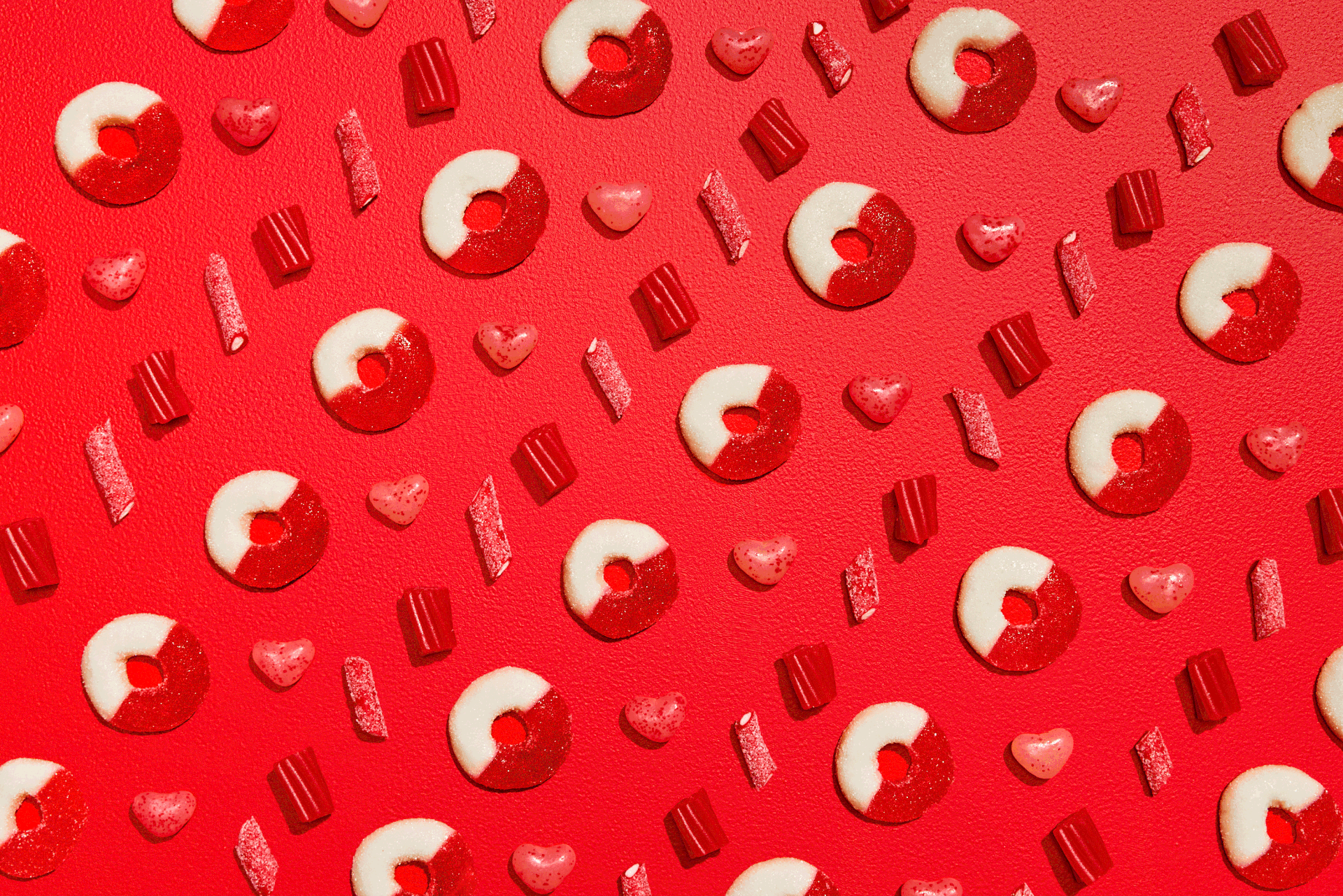 Pattern of decorated candies and candies in heart shapes on a red background, with some candies shaped like donuts with red and white icing.